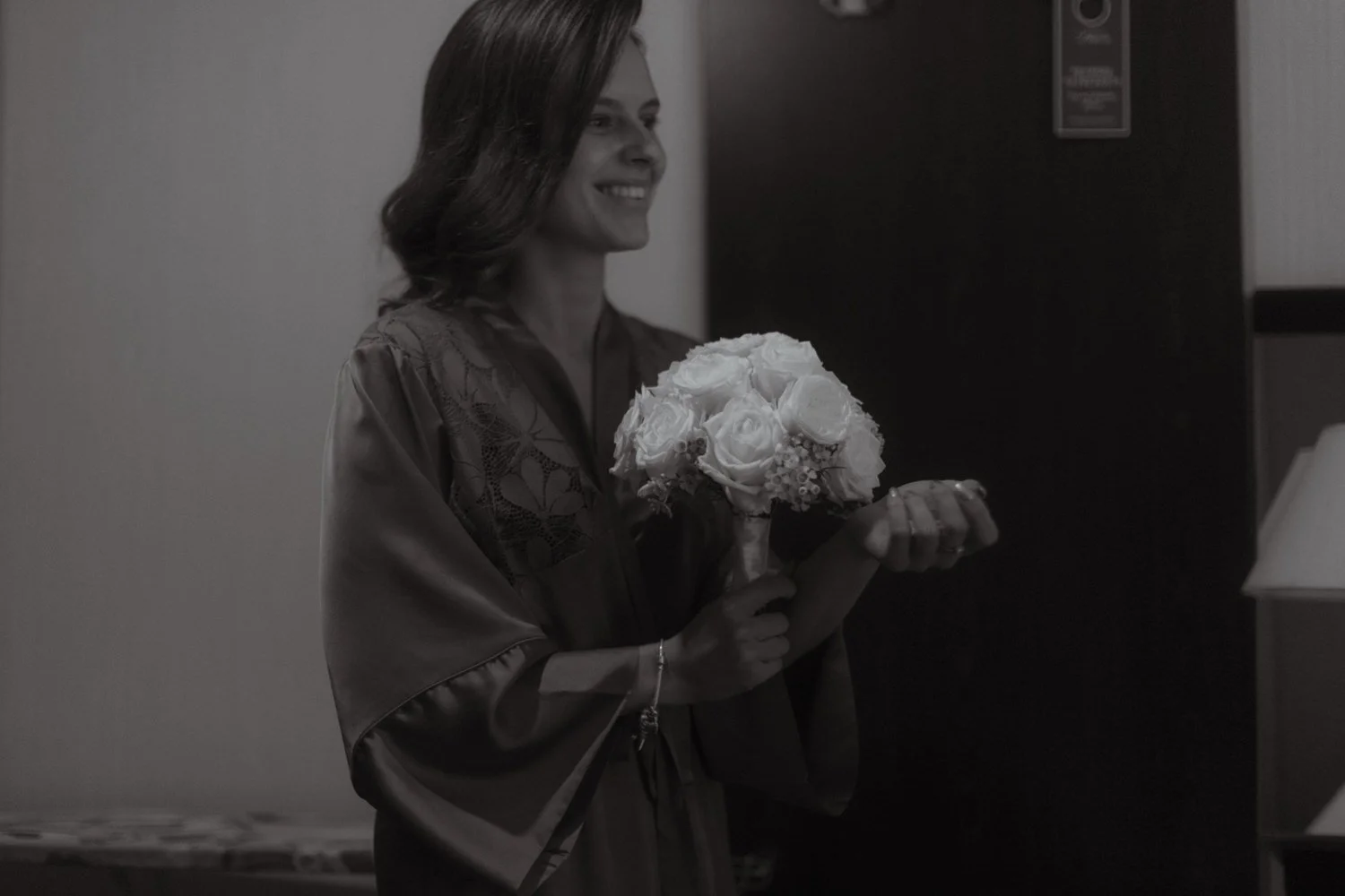 A woman in a satin robe is holding a bouquet of roses and smiling inside a room.