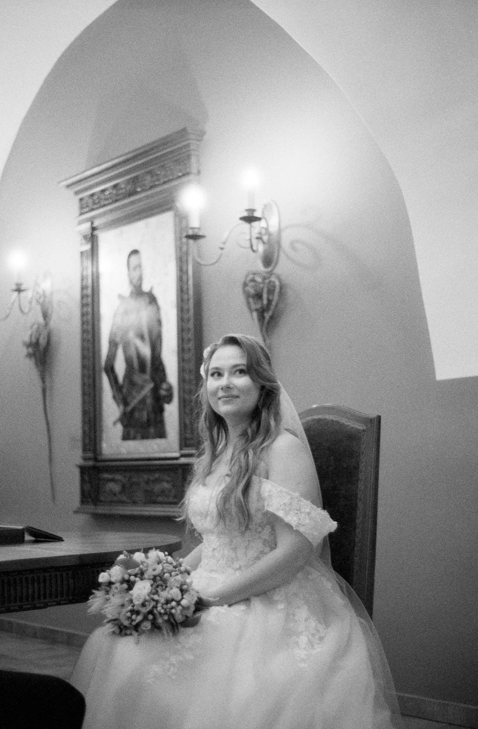 A woman in a wedding dress sitting and holding a bouquet in a decorated room, with a portrait and ornate wall sconces in the background.