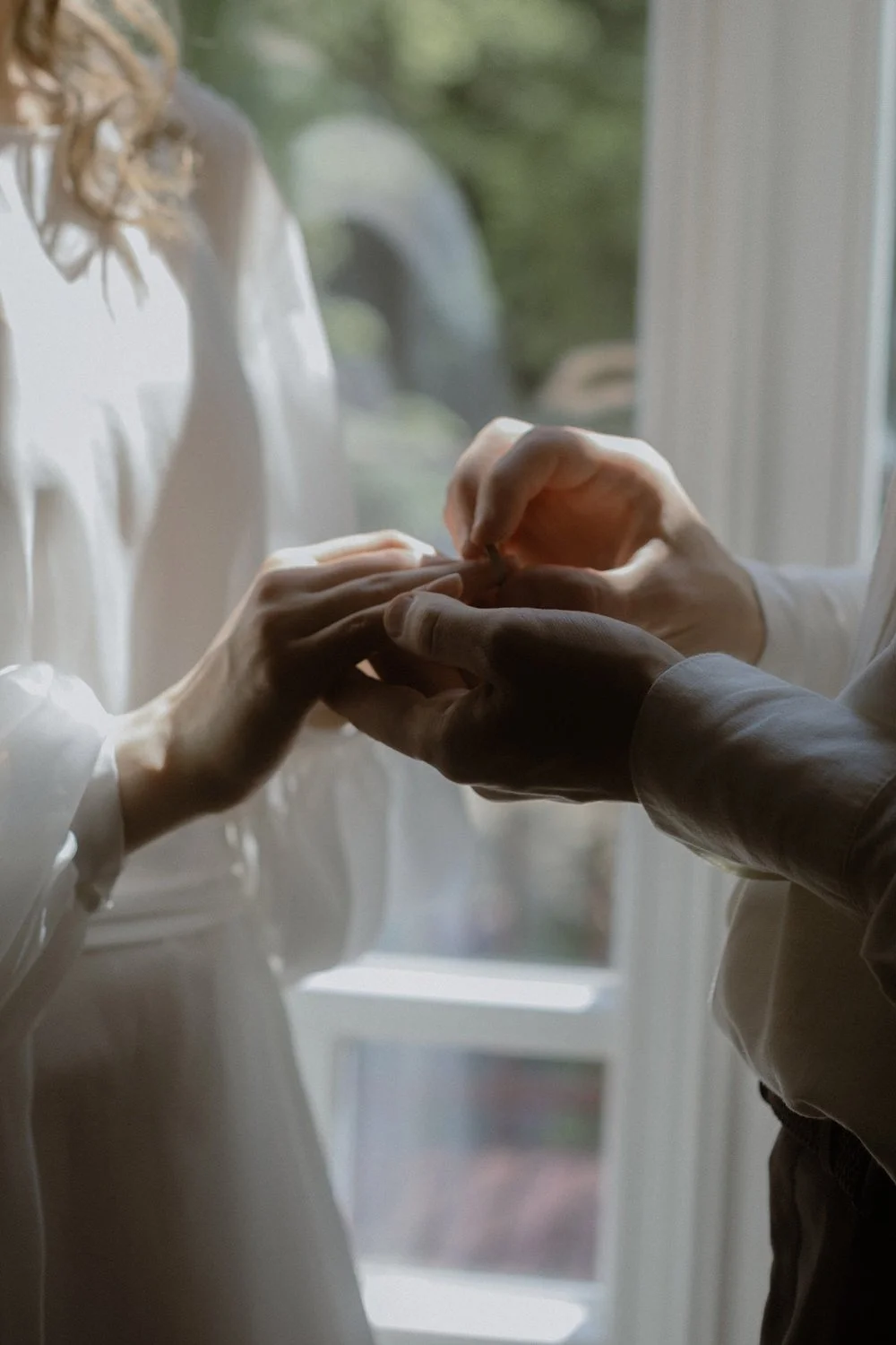 Close-up of two people holding hands, with one person putting a ring on the other's finger near a window with natural light.