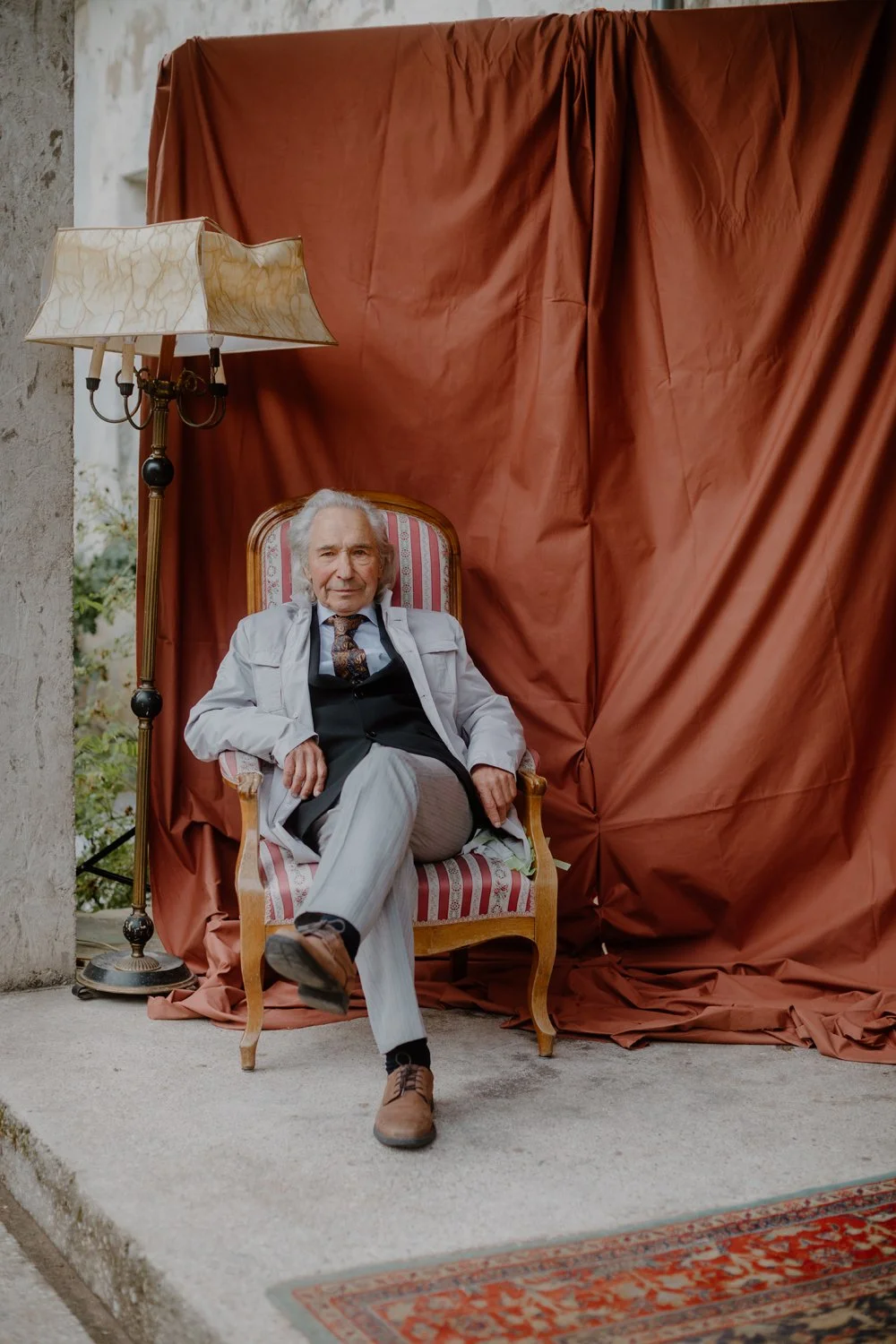 An elderly man, grooms father, with white hair sits in a vintage upholstered armchair with red and white striped fabric. He wears a light gray suit, black vest, patterned tie, and tan shoes. A tall vintage standing lamp with a cream-colored, marbled 