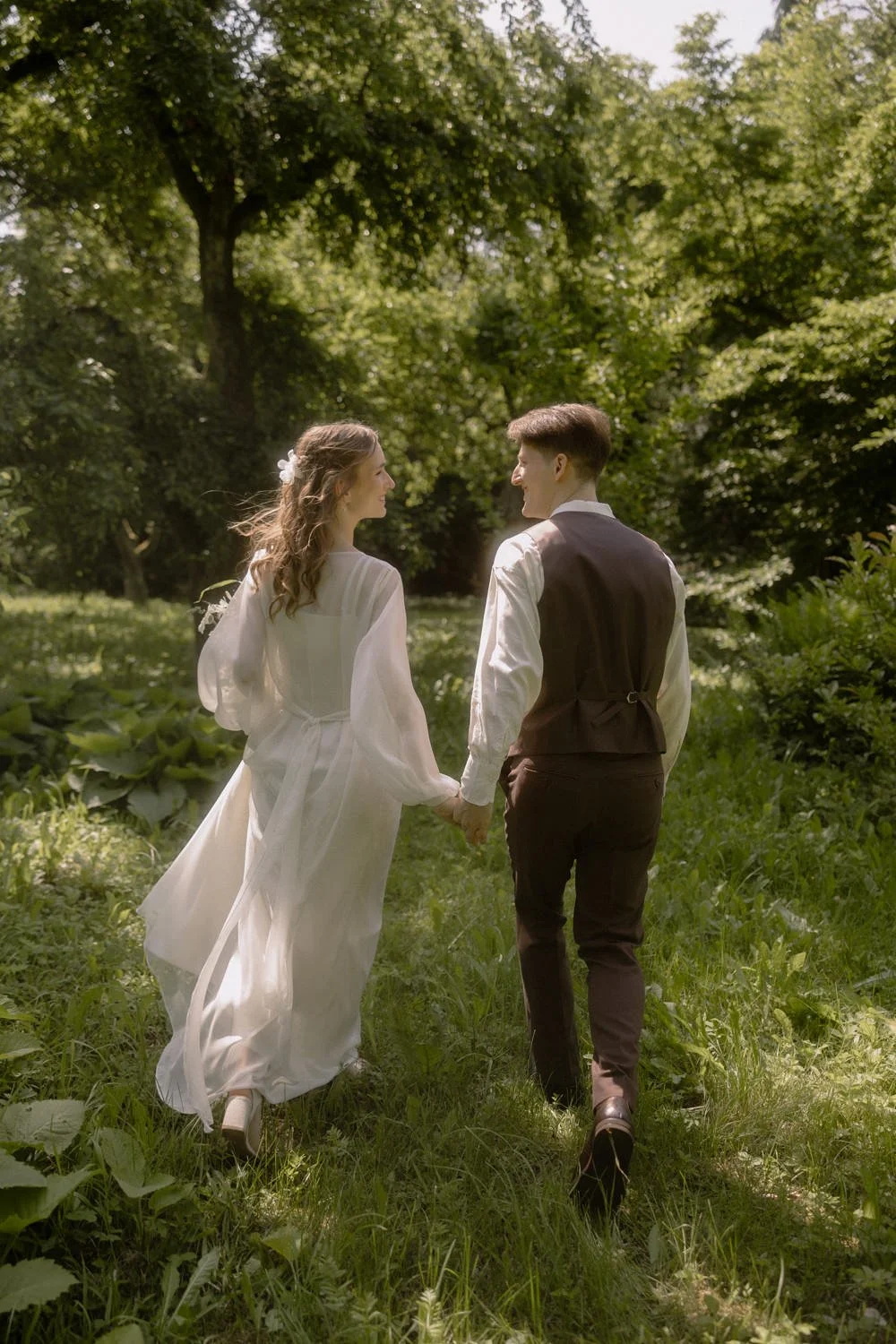 A couple in vintage wedding attire holding hands and walking through a lush green forest.
