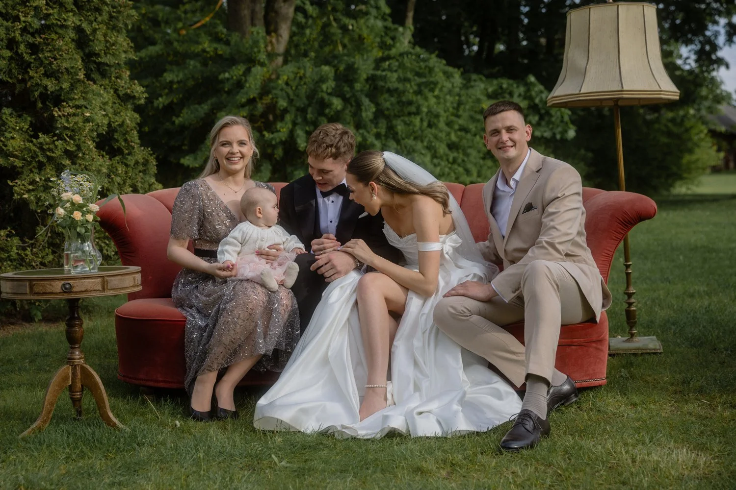 Group of six people sitting on a red sofa outdoors, including a bride in a white wedding dress, four adults, and a young girl holding a baby, all smiling.