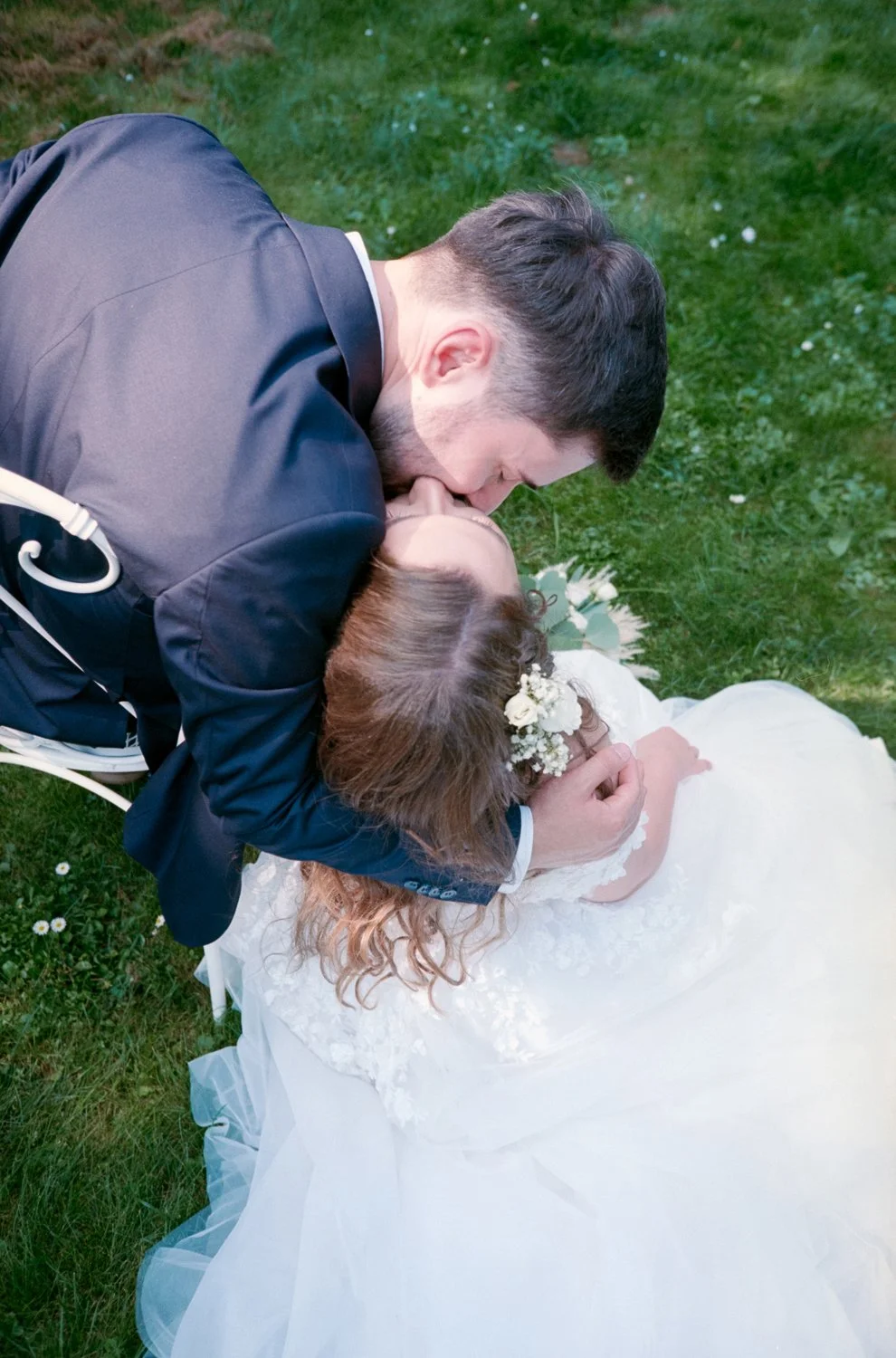 A man leaning over a woman in a wedding dress, passionately kissing her on the lips while she lies on the grass with her eyes closed, surrounded by greenery and small flowers.