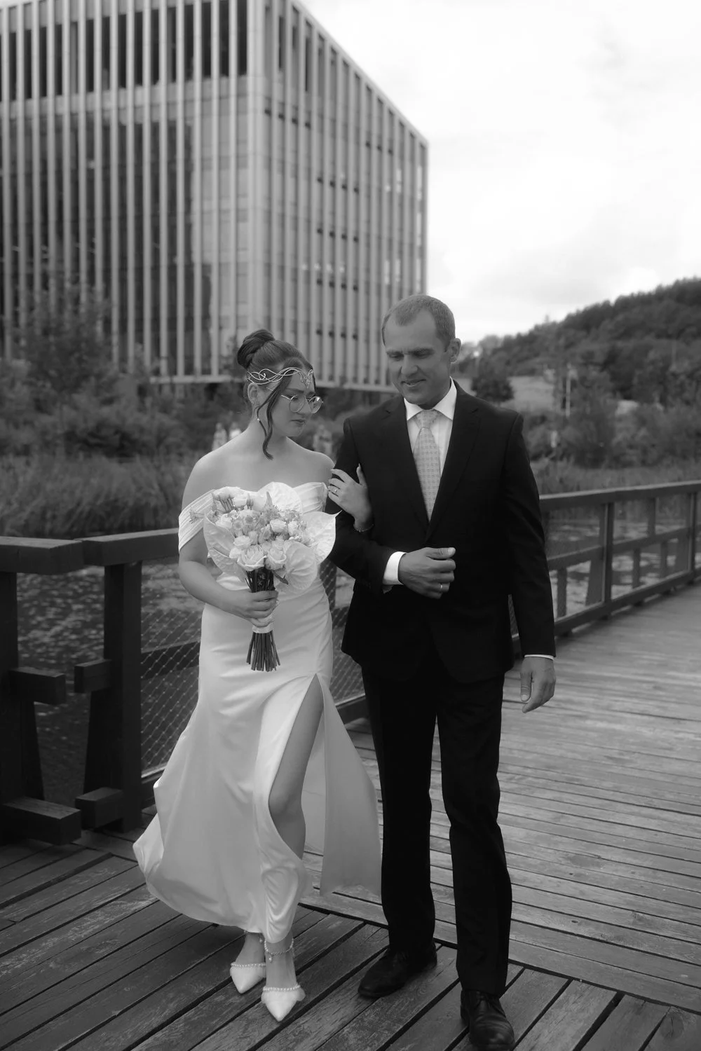 A bride in a white wedding dress and a groom in a black suit walk arm in arm on a wooden bridge, with a modern office building and trees in the background.