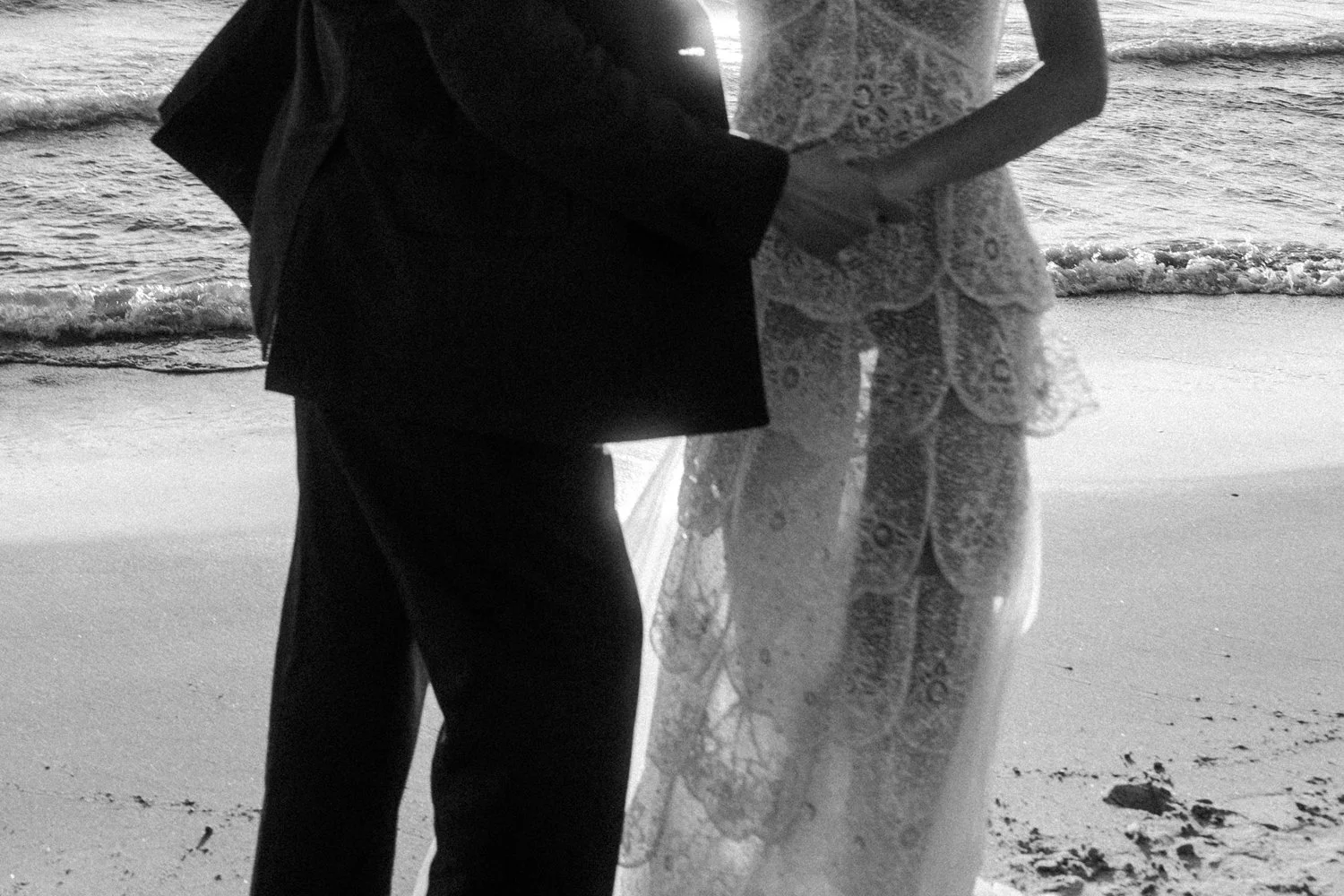 A couple holding hands on the beach, with the ocean and waves in the background, in black and white.