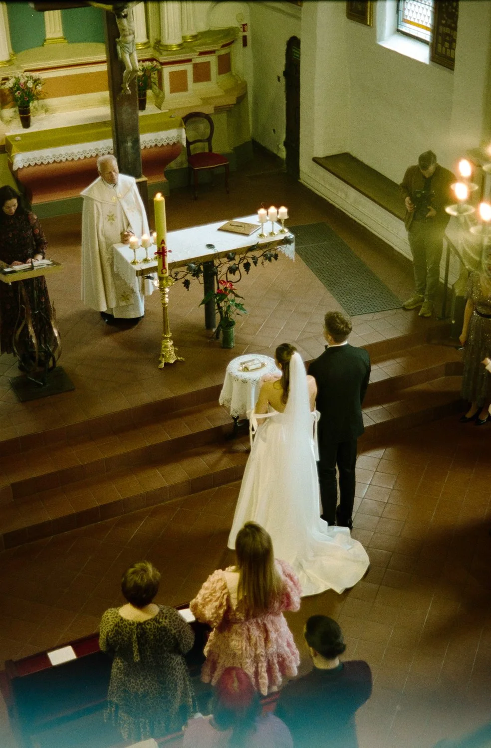 A church wedding ceremony taking place with a bride and groom standing in front of a priest at the altar. The bride is in a white wedding dress with a veil and the groom is in a dark suit. Several guests are present, some seated and some standing, wa