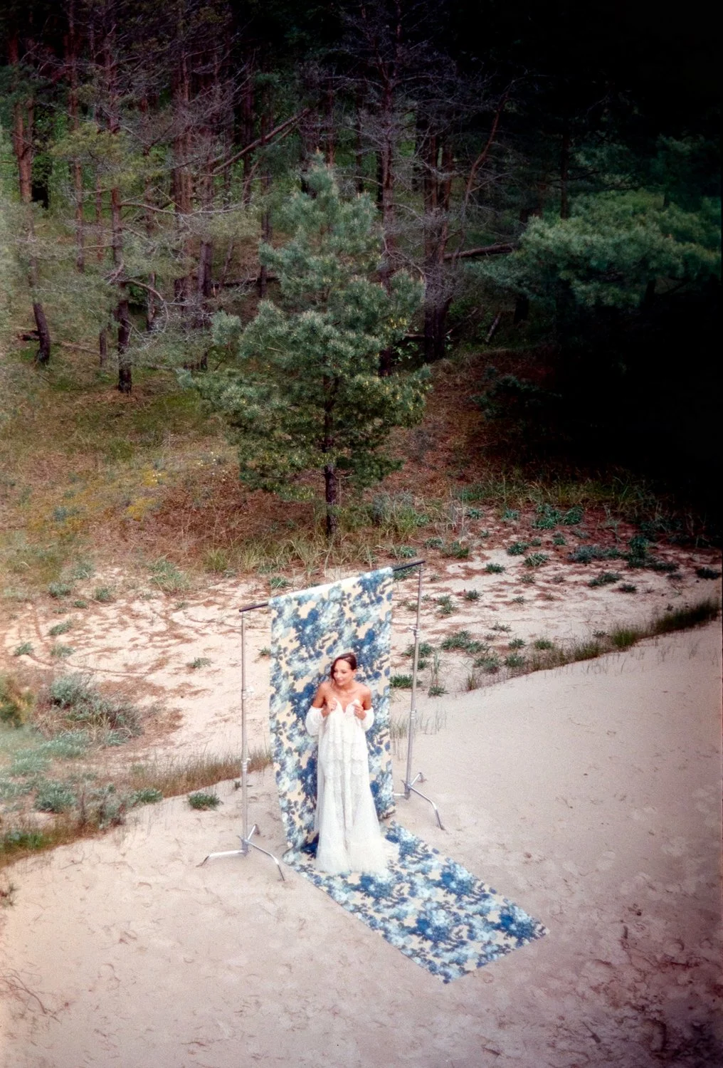A woman in a white dress standing on a patterned backdrop and runner at outdoor photoshoot in a sandy, wooded area.