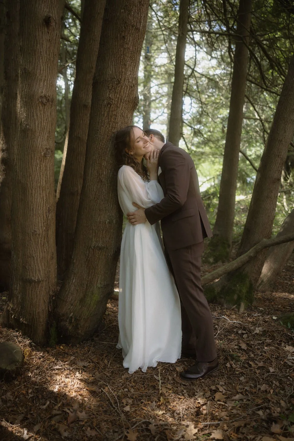 A bride and groom sharing a tender moment in a wooded area, standing close to a tree, with sunlight filtering through the leaves.