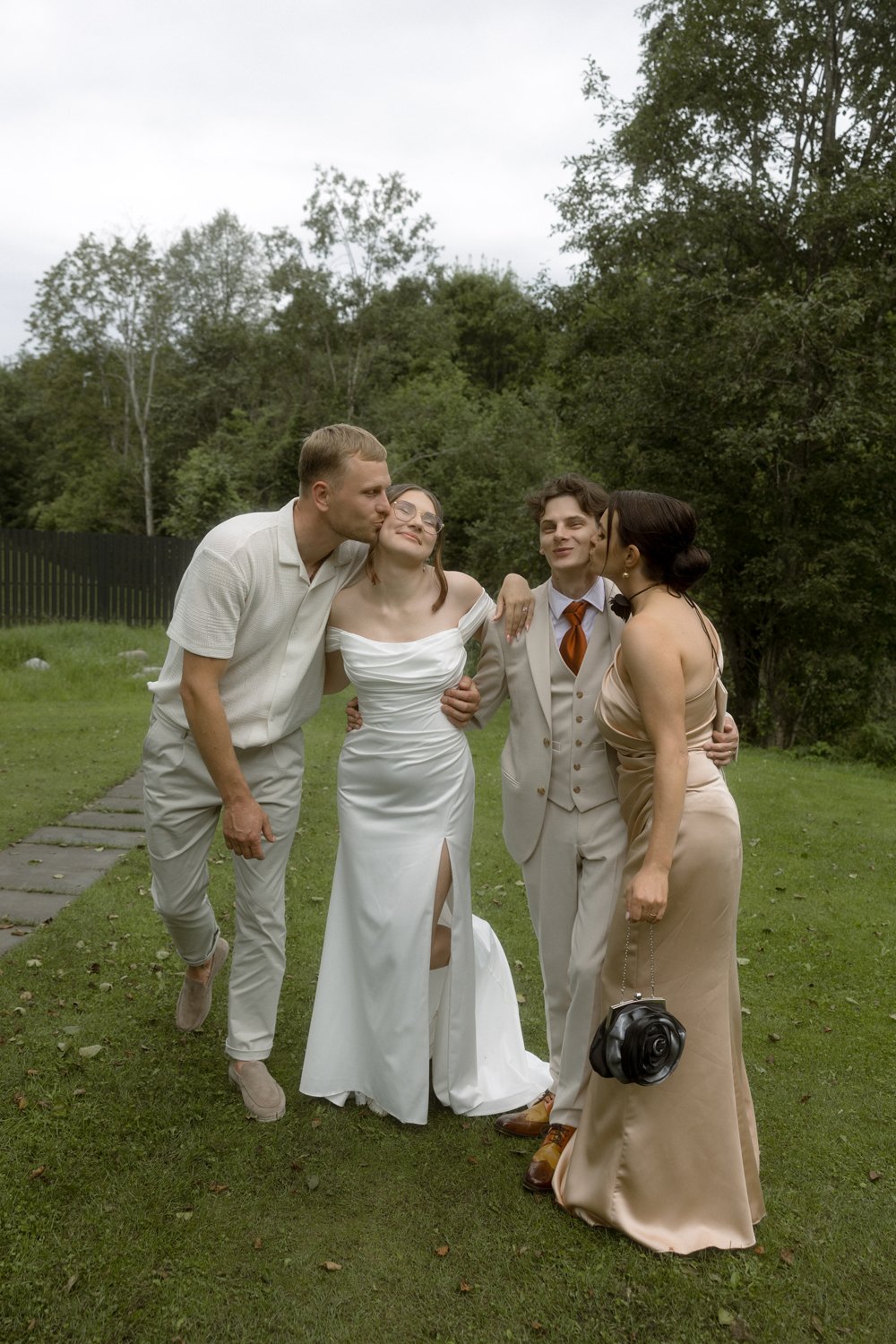Four people at a wedding, three men and a woman, standing outdoors on a grassy area, with trees and shrubs in the background, dressed in formal attire, with some kissing and one holding a camera.