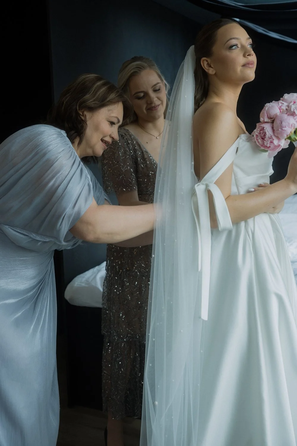 A bride in a white wedding dress holding a bouquet of pink peonies, standing with two women adjusting her dress, one in a gray dress and the other in a sparkly dress, in a dimly lit room.