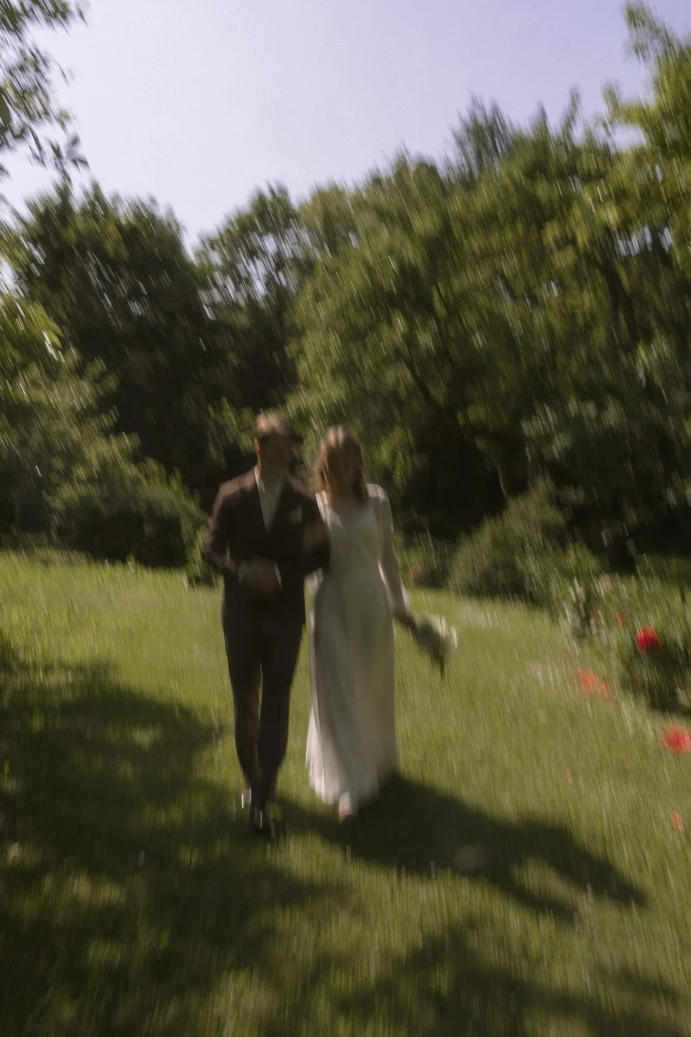 A blurred photo of a couple walking outdoors on a sunny day, with trees and grass in the background. The woman is wearing a white dress holding a bouquet, and the man is in a dark suit.