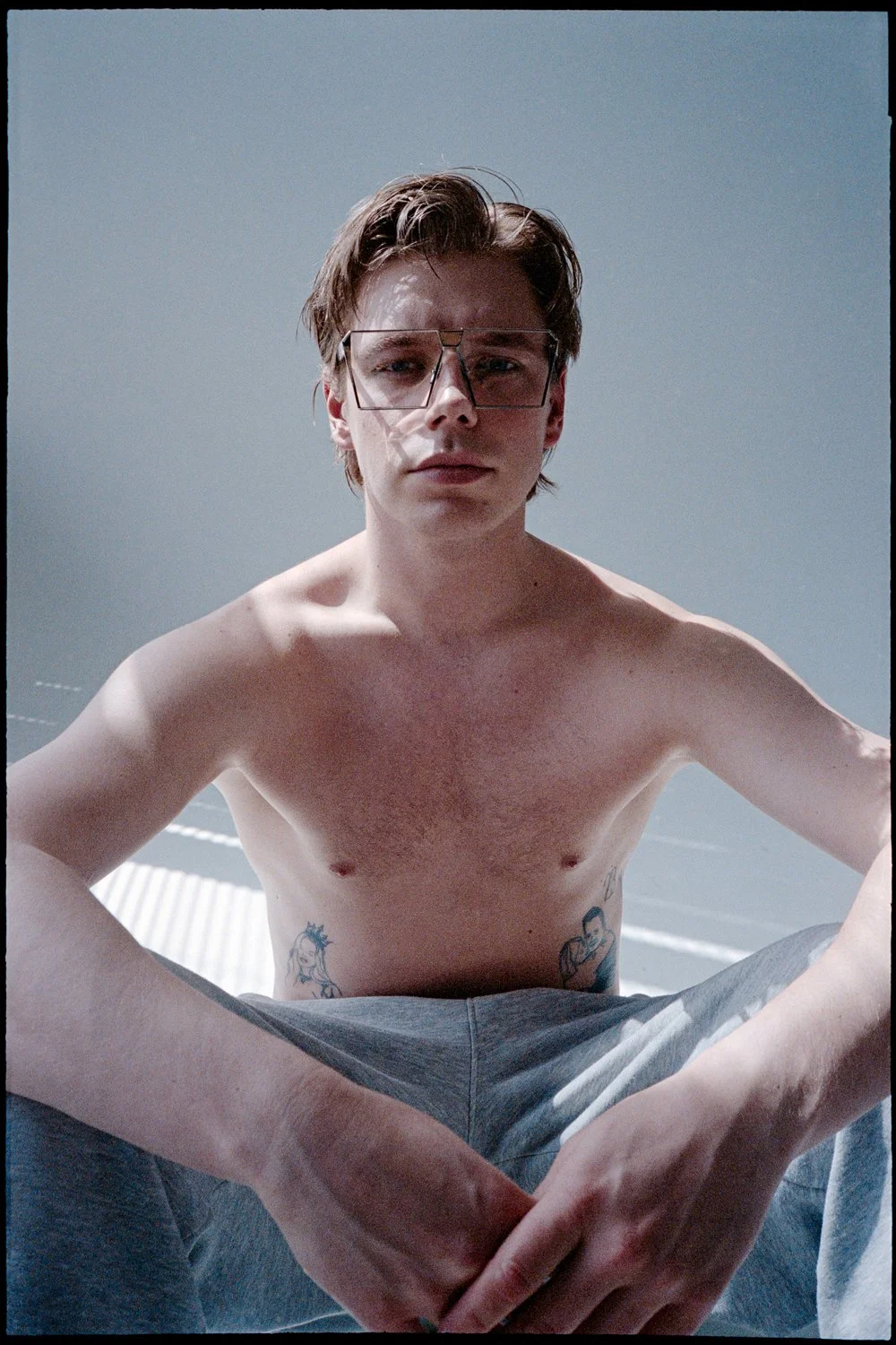 A young man with short brown hair and glasses is sitting shirtless against a plain, light-colored background. He has tattoos on his side and wears gray sweatpants.