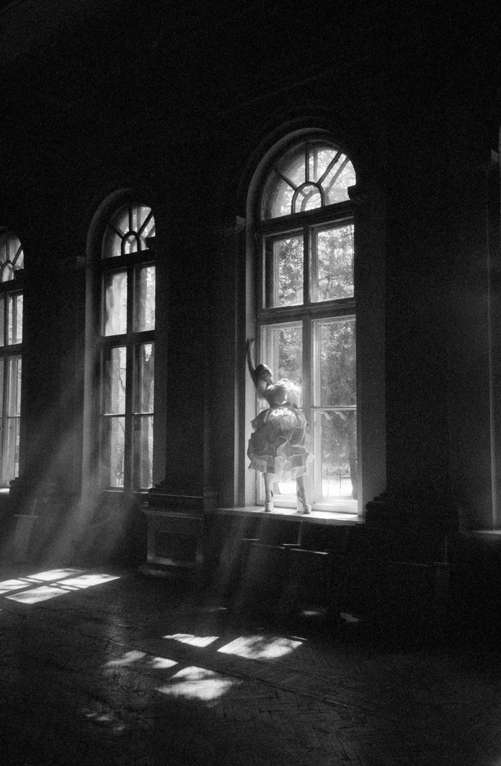 A black and white photo of a girl sitting on a windowsill in an old building, with sunlight streaming through large arched windows, casting shadows and light patterns on the floor.