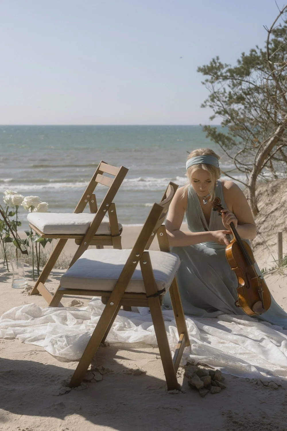 A woman sitting on the sand at the beach playing the violin, with two wooden chairs and white cushions nearby, and the ocean waves in the background.