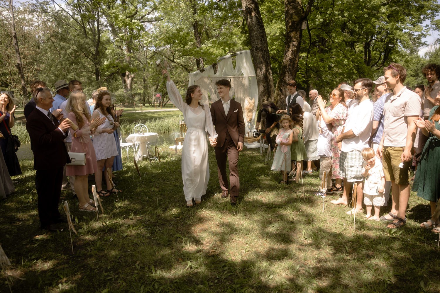 A bride and groom walking hand-in-hand down an outdoor aisle surrounded by family and friends at a garden wedding, with trees and grass in the background.