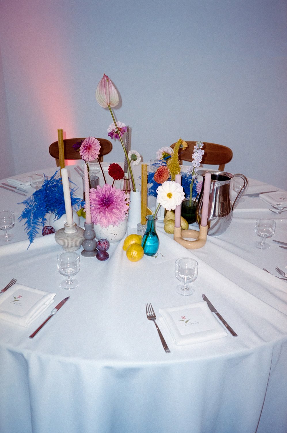 Elegant round table set for a meal with white tablecloth, assorted colorful flowers in vases and candles, and place settings with napkins, forks, knives, and small glasses, featuring a modern, floral theme.