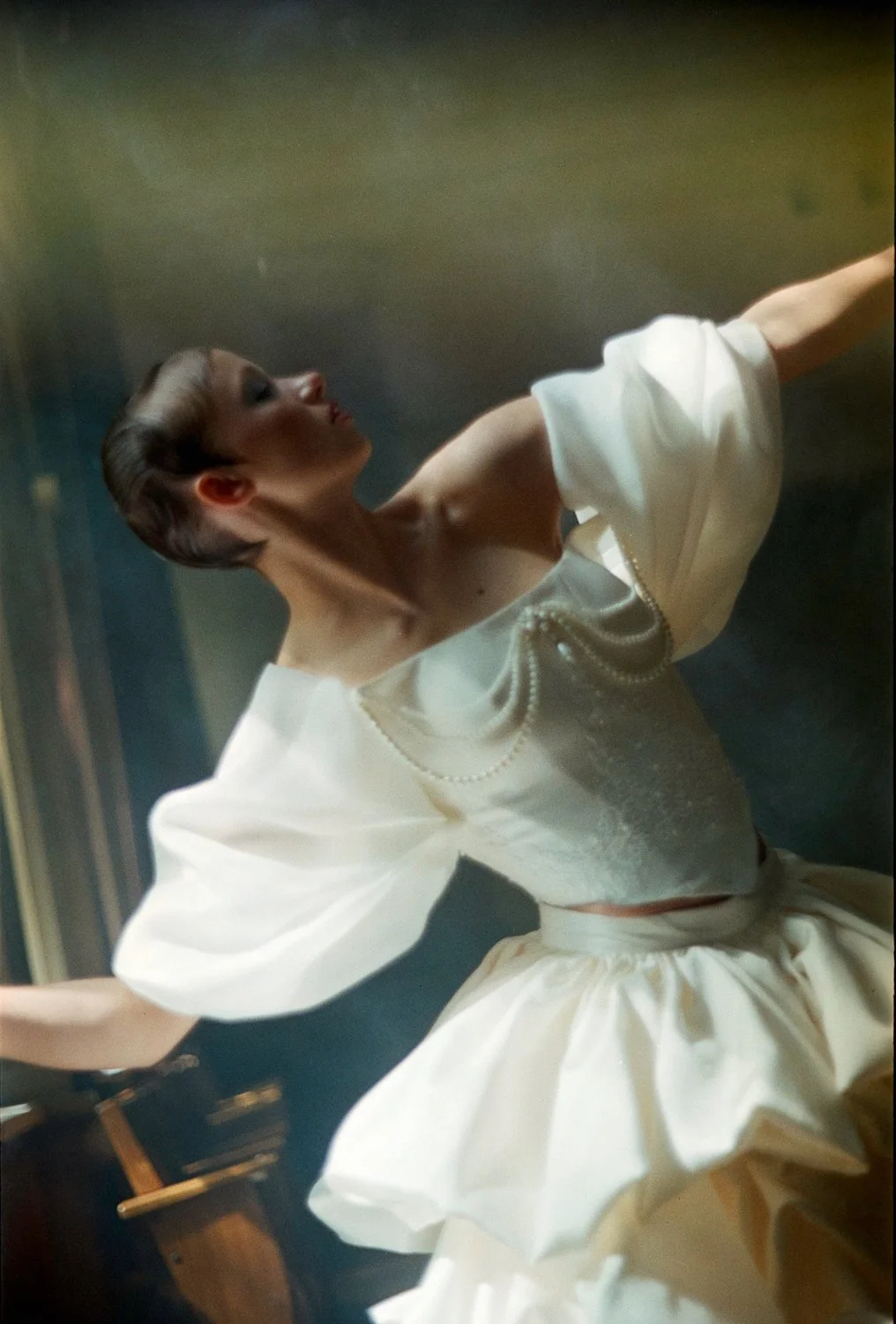 A woman in a vintage white dress with puffy sleeves and pearl details, posing sideways with her head turned upward and arms extended, in an artful, softly lit setting.