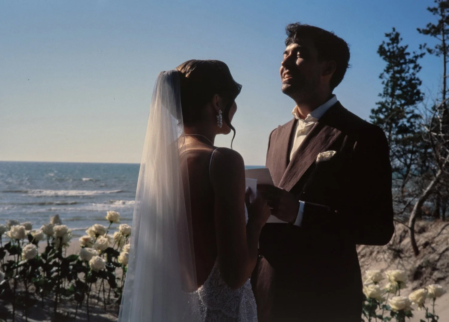 A couple getting married on the beach, with the ocean and trees in the background. The bride is wearing a veil and dress, and the groom is in a suit. They are holding hands and facing each other during the ceremony.