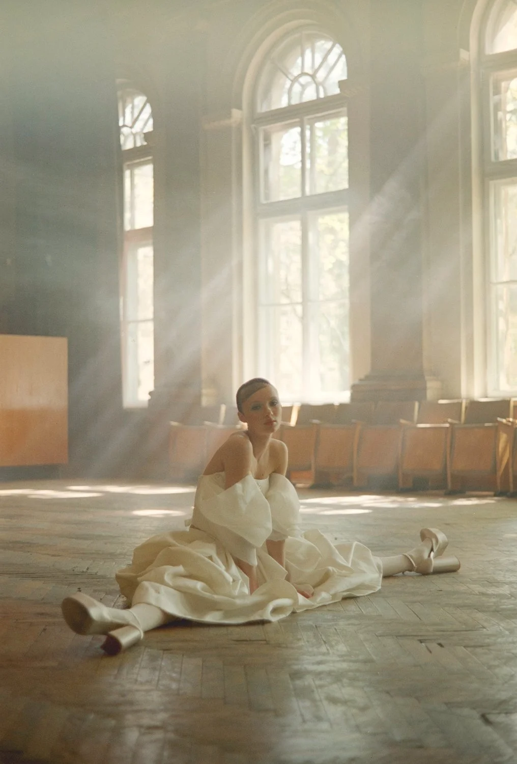 A woman in a white dress and high heels sits on the wooden floor of a large, sunlit room with tall windows and wooden chairs along the walls.