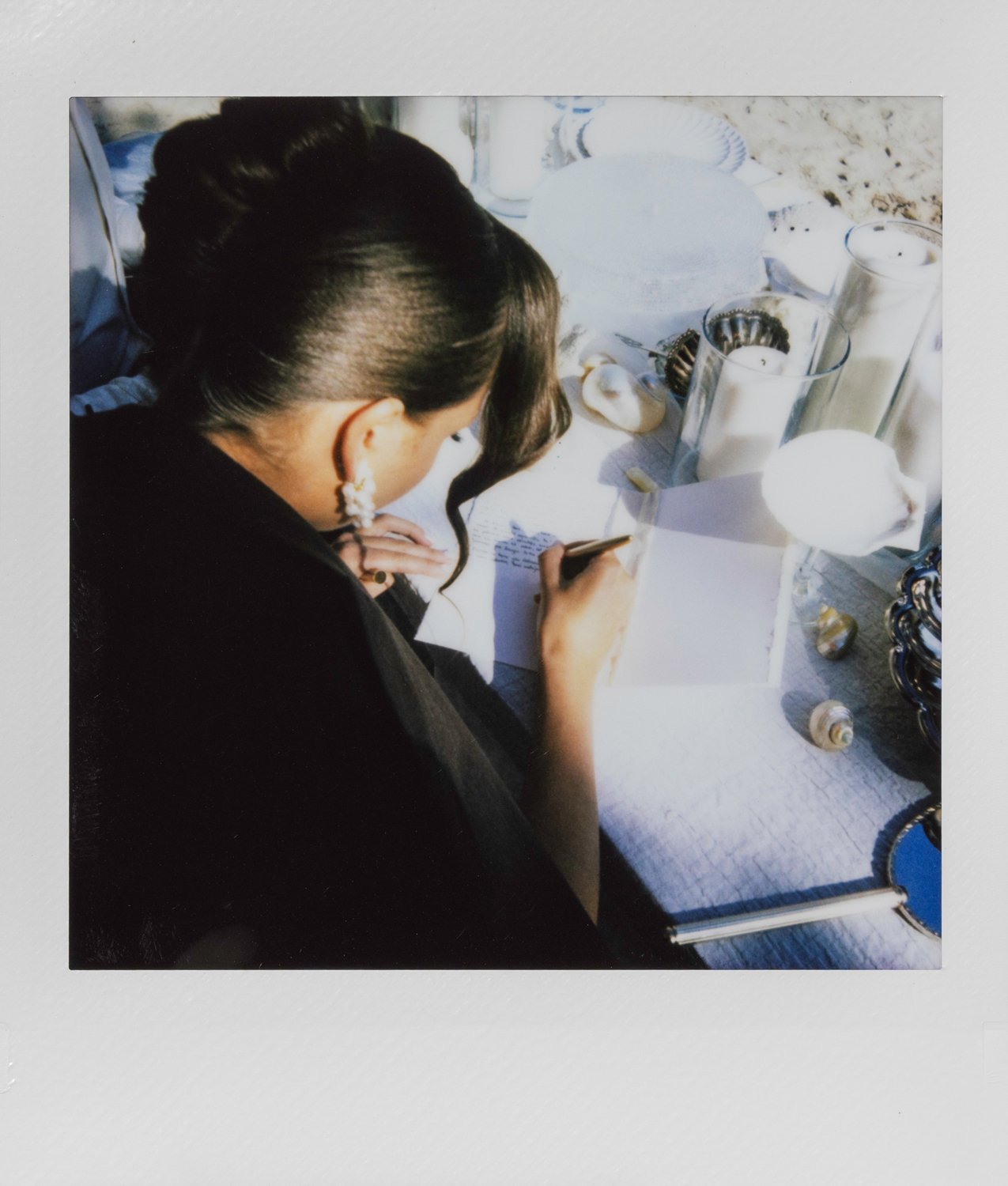 A woman with dark hair styled in an elegant updo, wearing earrings, writing on a piece of paper at a table set with white candles and decorative shells.