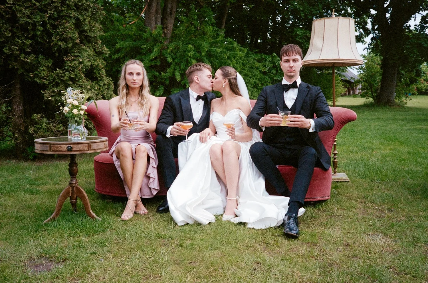 A bride and groom sitting on a vintage red sofa outside, with two friends, all dressed formally for a wedding. The bride is kissing the groom while two friends sit beside them holding drinks, one woman with a pink dress on the left, and a man in a bl