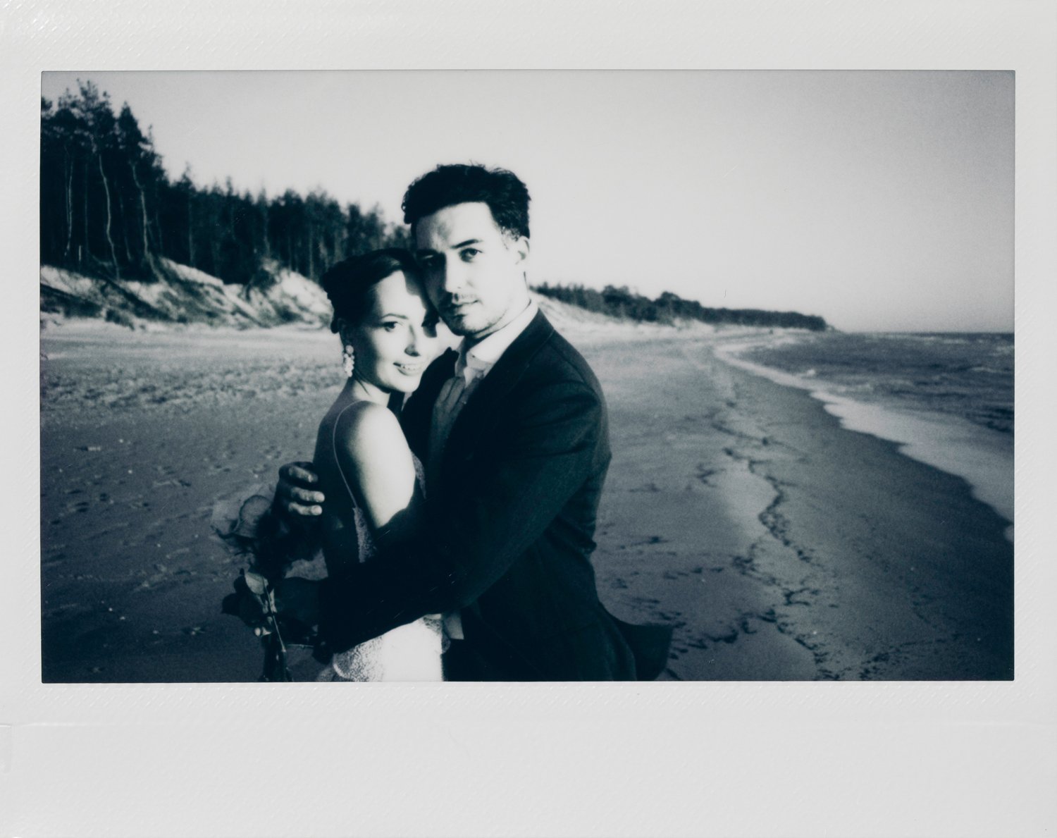Black and white photo of a couple embracing on a beach, with ocean waves and trees in the background.
