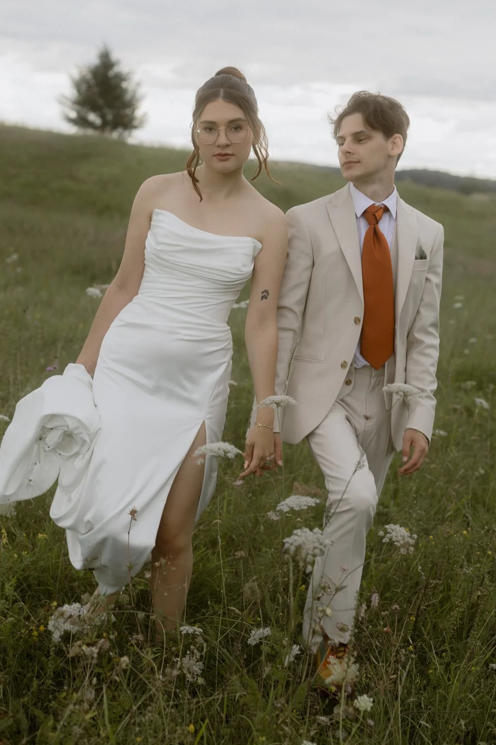 A woman in a white wedding gown and a man in a beige suit walk hand in hand through a field of grass and wildflowers, with a cloudy sky in the background.