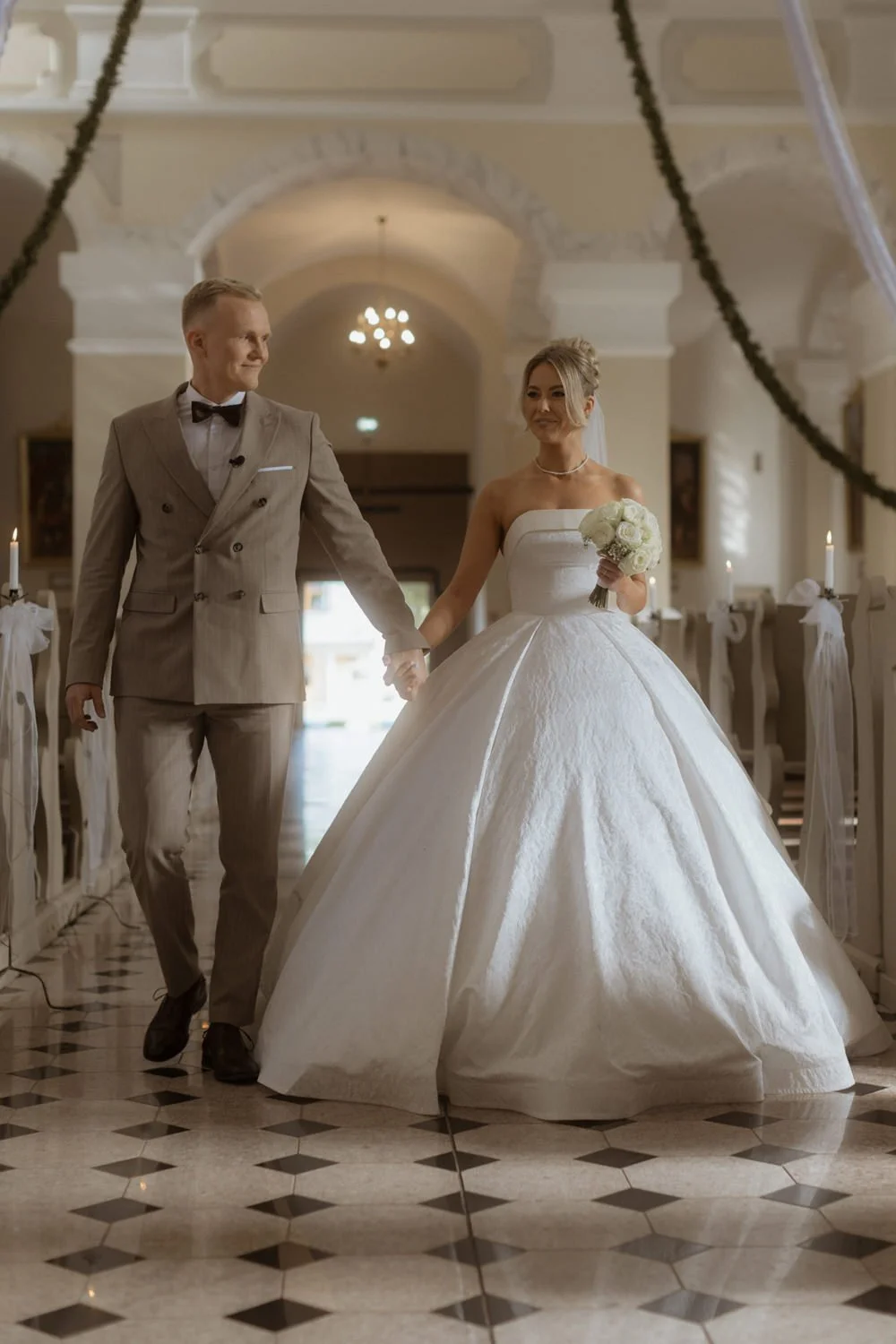 A bride and groom walking hand in hand down the aisle inside a church decorated with candles and flowers. The bride is wearing a strapless white wedding gown and holding a bouquet of white flowers, while the groom is dressed in a light gray suit with