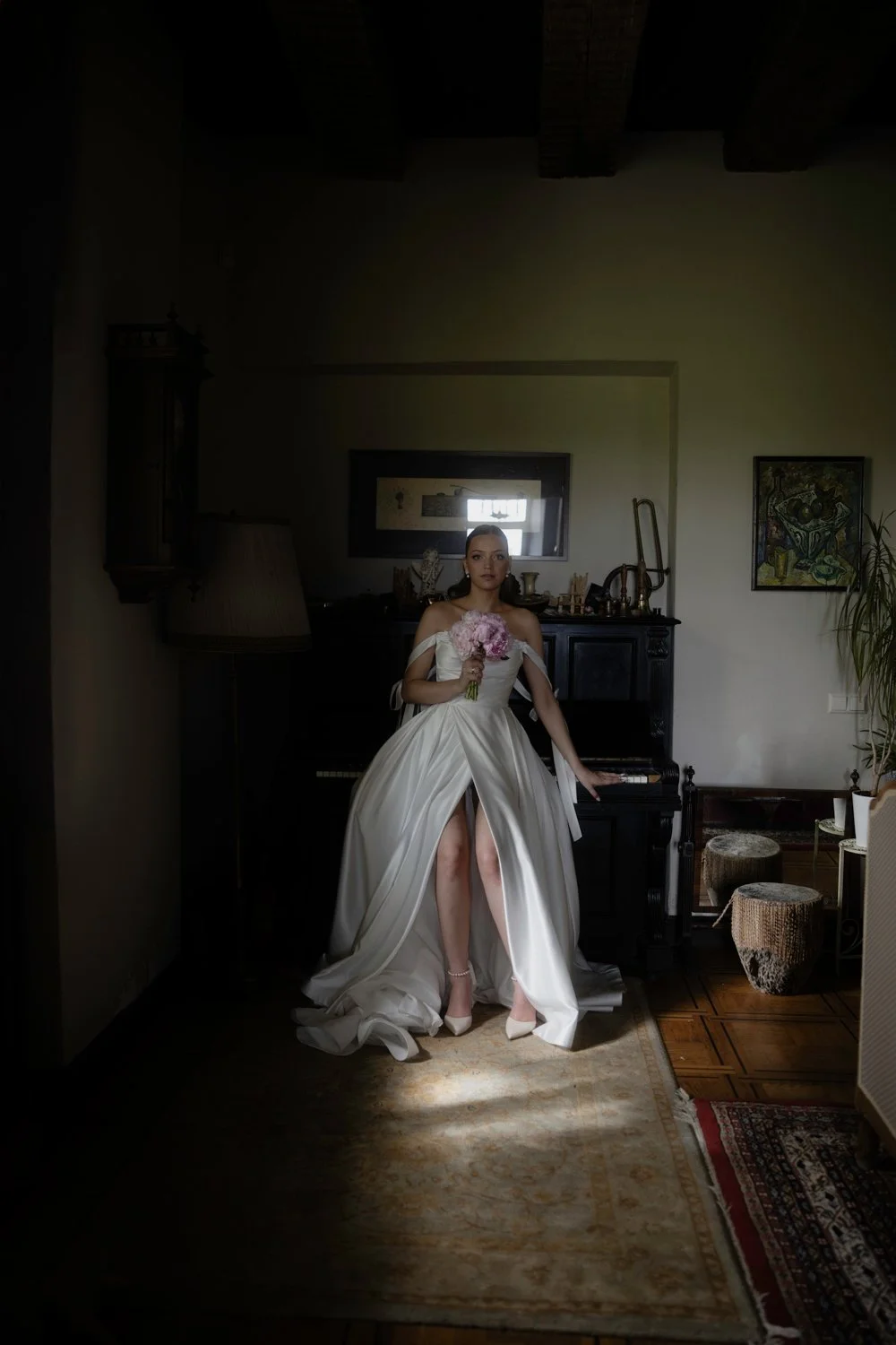 A woman in a wedding dress holding a pink bouquet sitting next to a black piano in a dimly lit room.