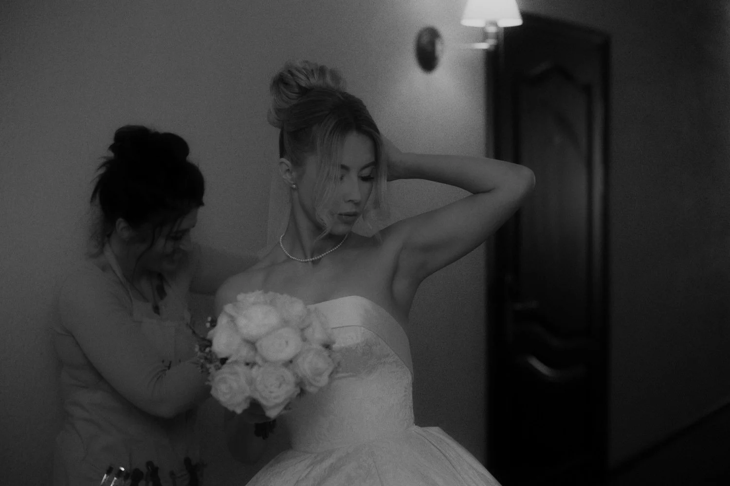 A woman in a wedding dress holds a bouquet of roses while another woman helps adjust her dress in a dimly lit room.