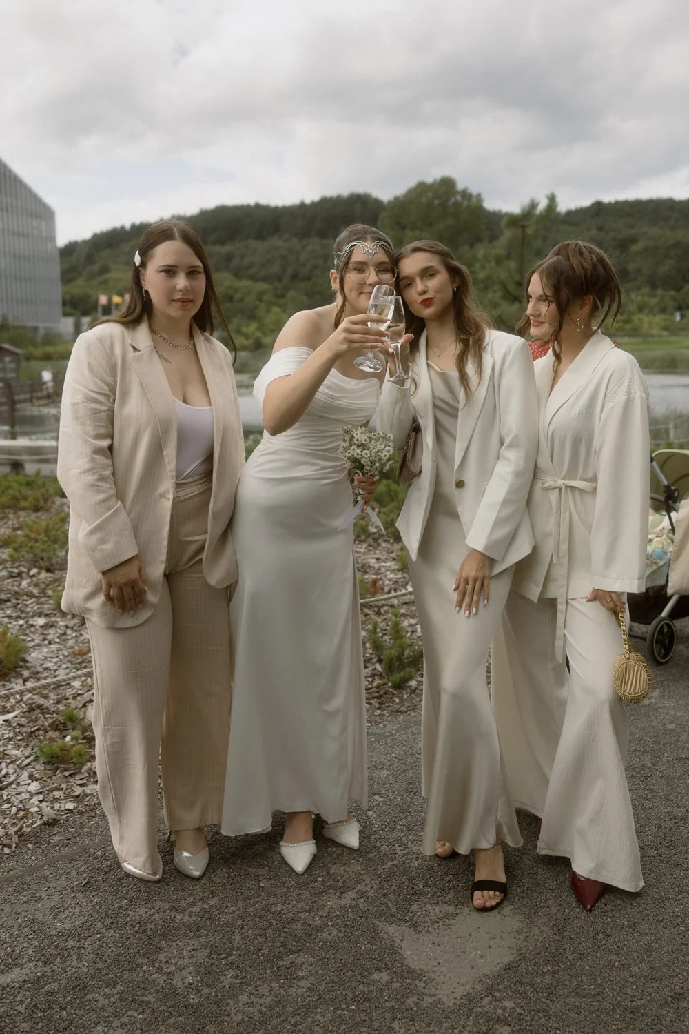 Group of four women celebrating outdoors, dressed in white and beige outfits, holding a glass of champagne, with a scenic background of trees and a lake.