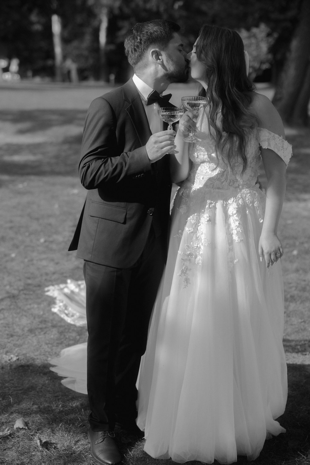 A black and white photo of a bride and groom sharing a kiss outdoors, holding champagne glasses. The groom is dressed in a tuxedo, and the bride wears a lace wedding gown with floral details.