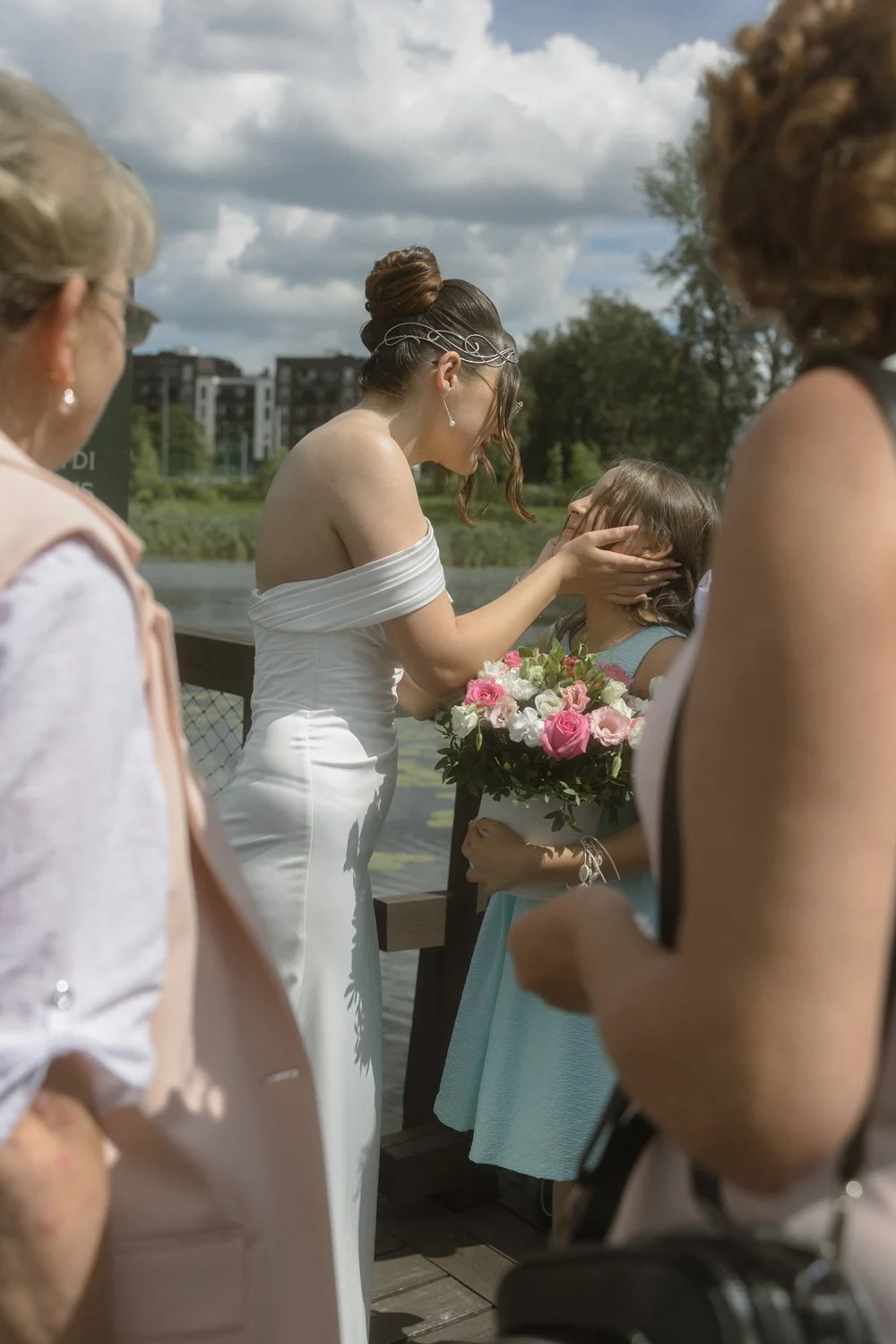 A bride with brown hair in an white off-shoulder wedding dress is holding a young girl with brown hair, who is holding a bouquet of pink and white roses, while gently touching the girl's face during an outdoor wedding ceremony by a lake.