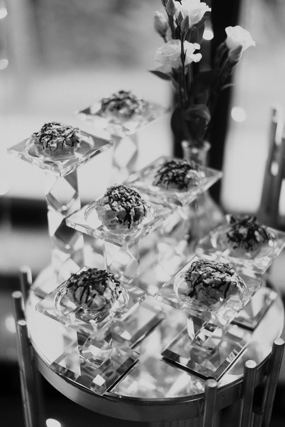 A black and white photo of a dessert display with small pastries topped with chocolate drizzle, placed on clear glass stands. In the background, there is a vase with flowers.