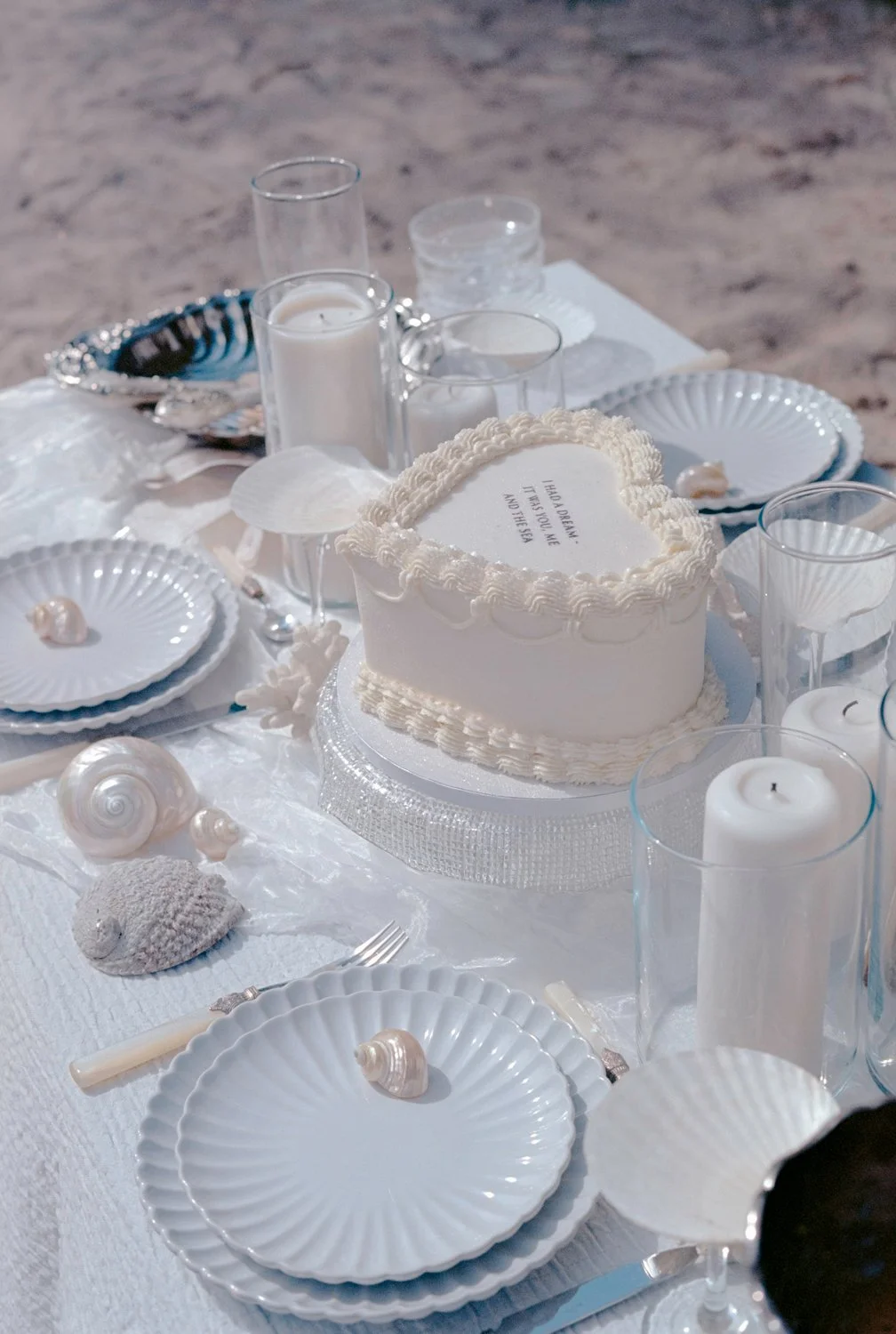 A beachside table set with white plates, shells, candles, and a heart-shaped cake with a message, overlooking sandy beach.