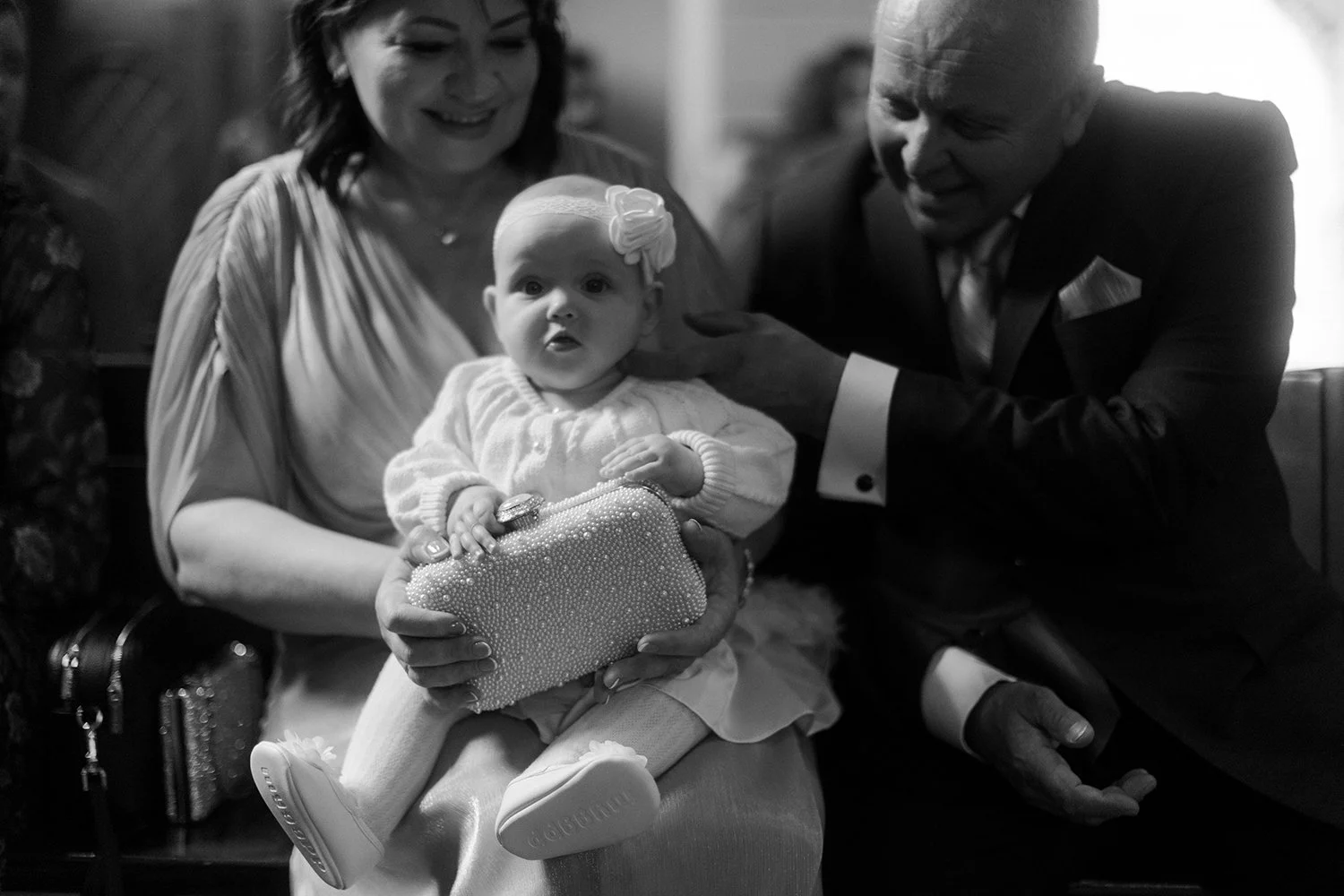 A baby girl sitting on a woman's lap, holding a pearl-embellished clutch with her left hand, with a man touching her shoulder. The woman is smiling, and the baby has a neutral expression, wearing a bow headband and a knitted outfit, in a black-and-wh