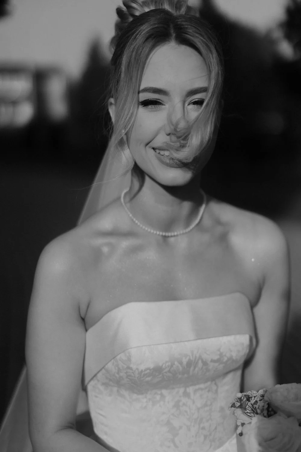 A smiling woman in a strapless dress, wearing a pearl necklace, with her hair styled and some of it falling over her face. She has a joyous expression, holding flowers, outdoors during twilight.