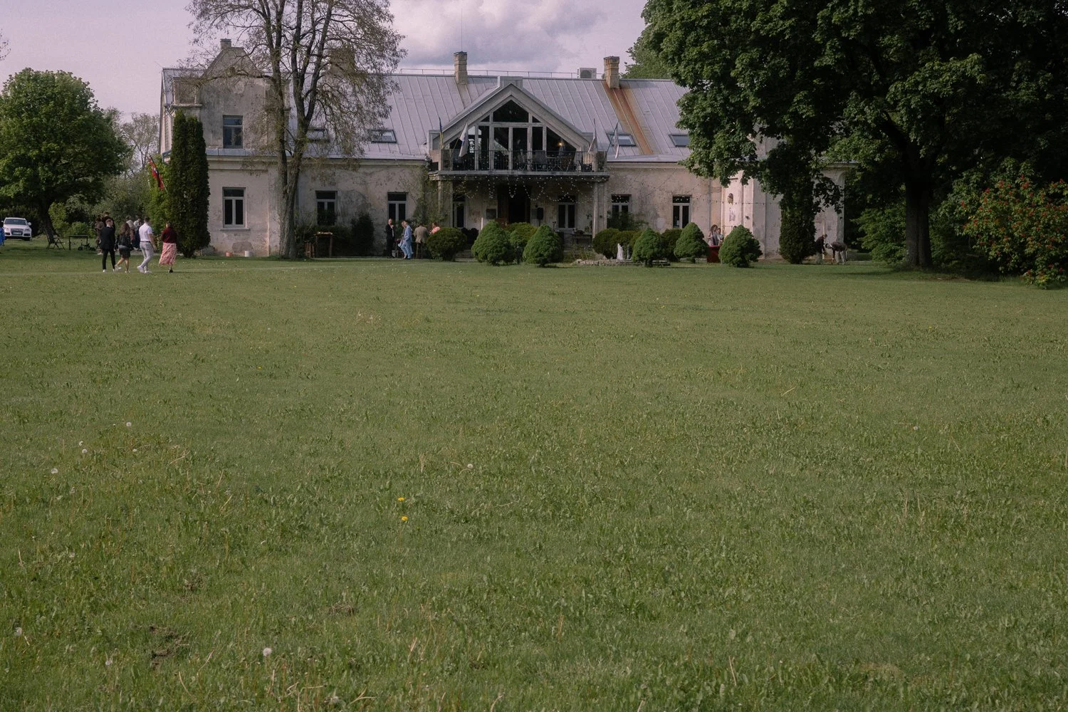 A large, old house with a metal roof, surrounded by a green lawn and tall trees. Several people are walking and standing near the house, and there are bushes and trees on either side of the house.