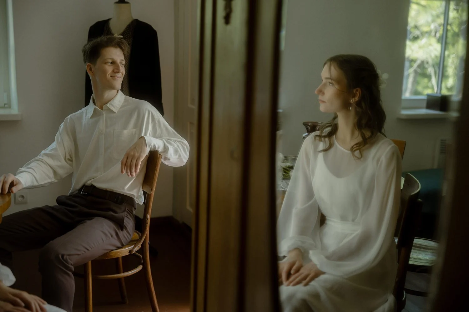 A young man and woman sitting at a table, engaged in conversation, in a softly lit room with natural light coming through a window. The woman is wearing a white dress, and the man is dressed in a white shirt and brown pants.