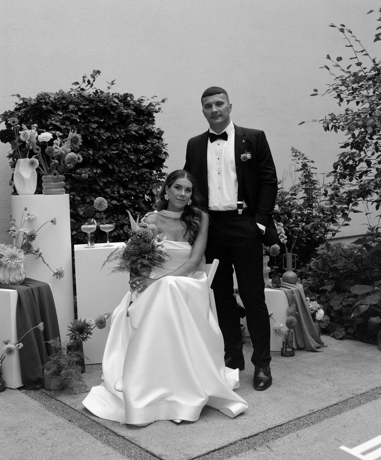 A bride in a wedding dress seated on a chair, holding a bouquet, with a groom in a tuxedo standing beside her, in a decorated outdoor setting with flowers and vases.