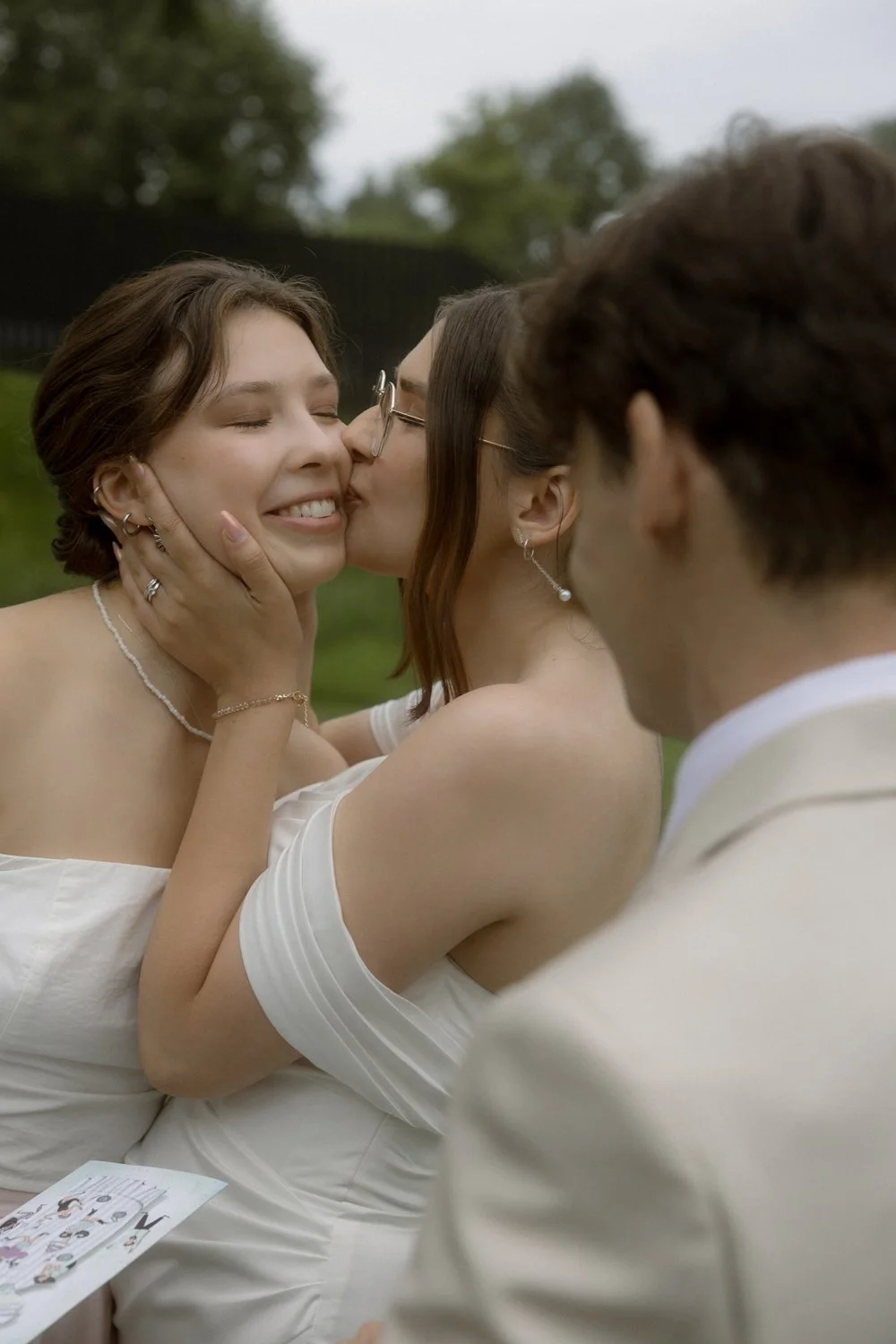 Two women sharing a kiss, with one touching the other's face, during a wedding ceremony. A man in a light-colored suit is also visible, observing the moment.