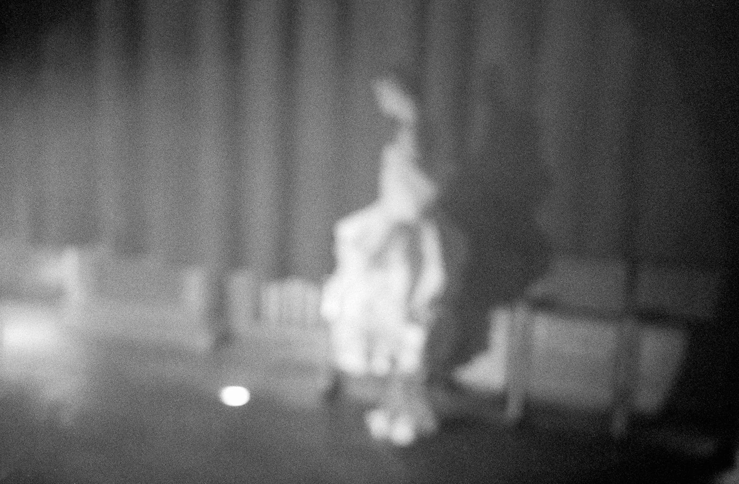 Blurry black and white photo of a person walking on a bridge at night, with trees and a railing visible.