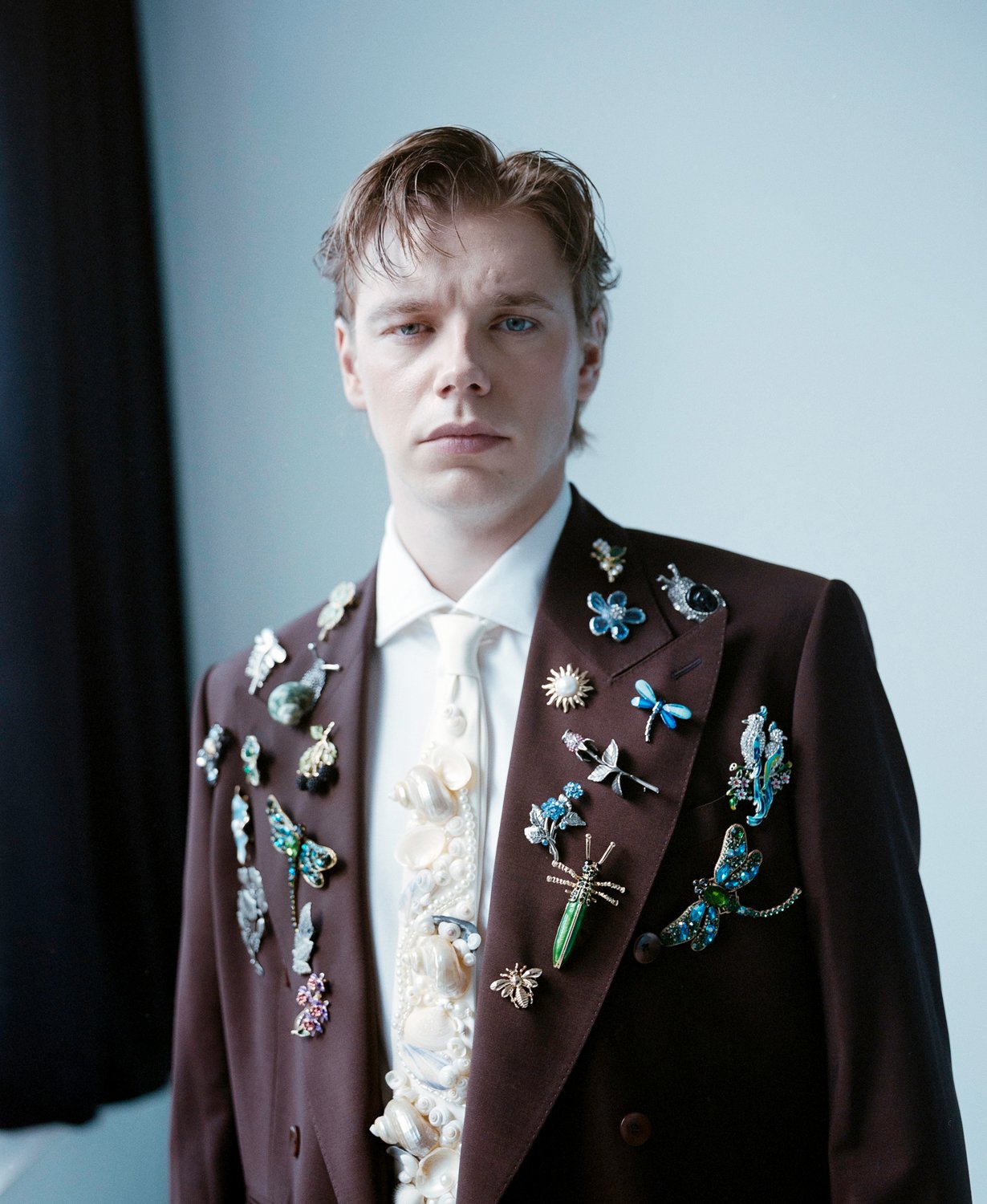 A young man with fair skin and light brown hair styled in a messy manner, wearing a dark brown suit jacket adorned with various decorative pins, a white dress shirt with a ruffled front, and a white tie, standing against a plain light-colored backgro
