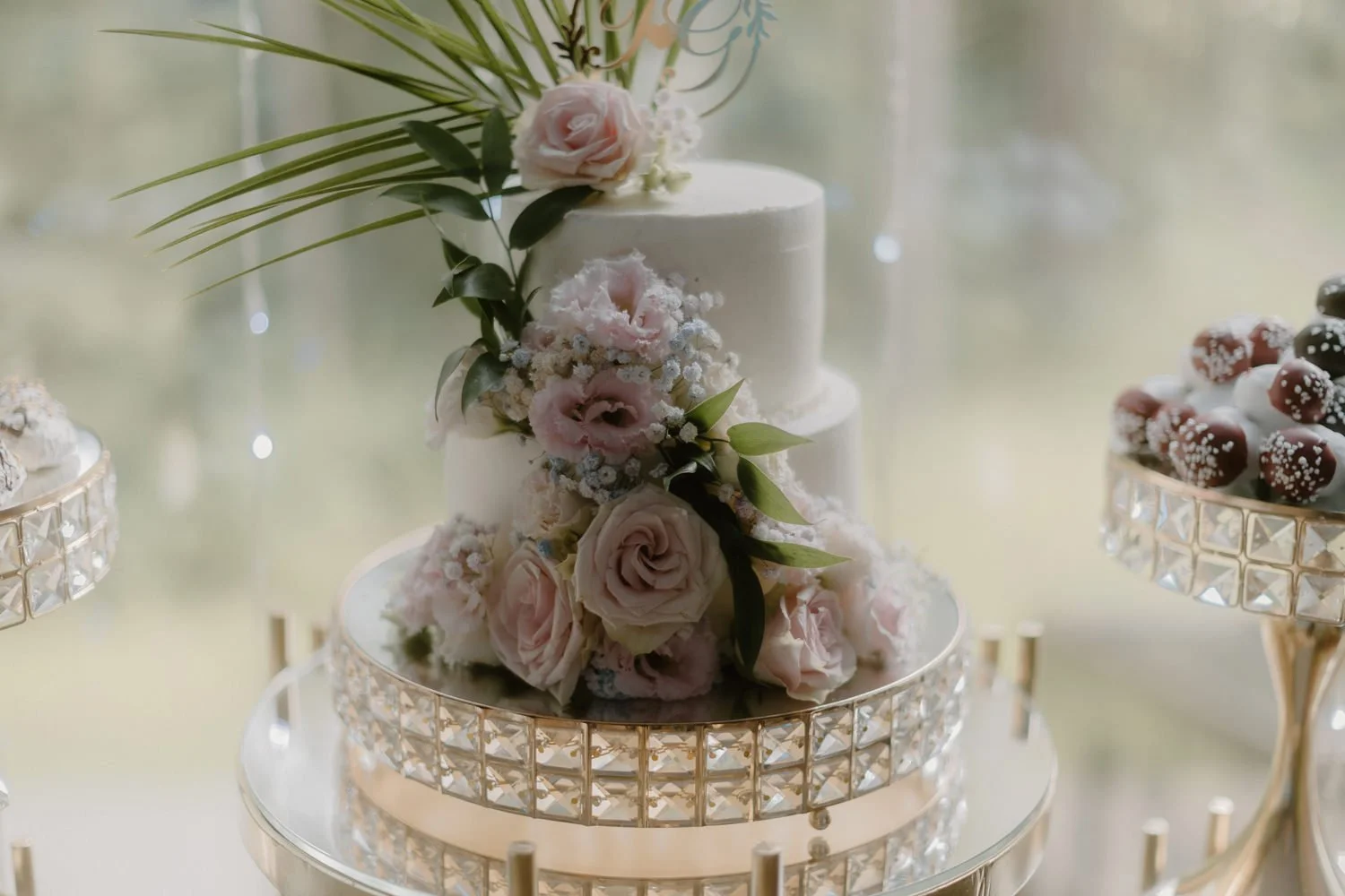 Elegant white wedding cake decorated with pink and white roses and greenery, displayed on a mirrored stand with crystal details.
