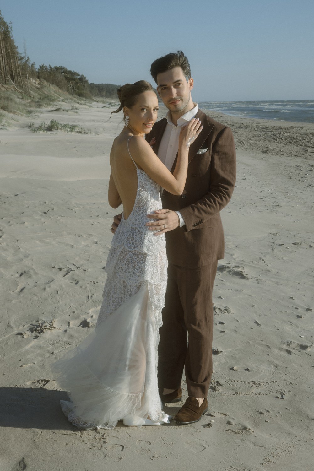 A bride and groom standing on a sandy beach, embracing each other in wedding attire, with the ocean and shoreline in the background.