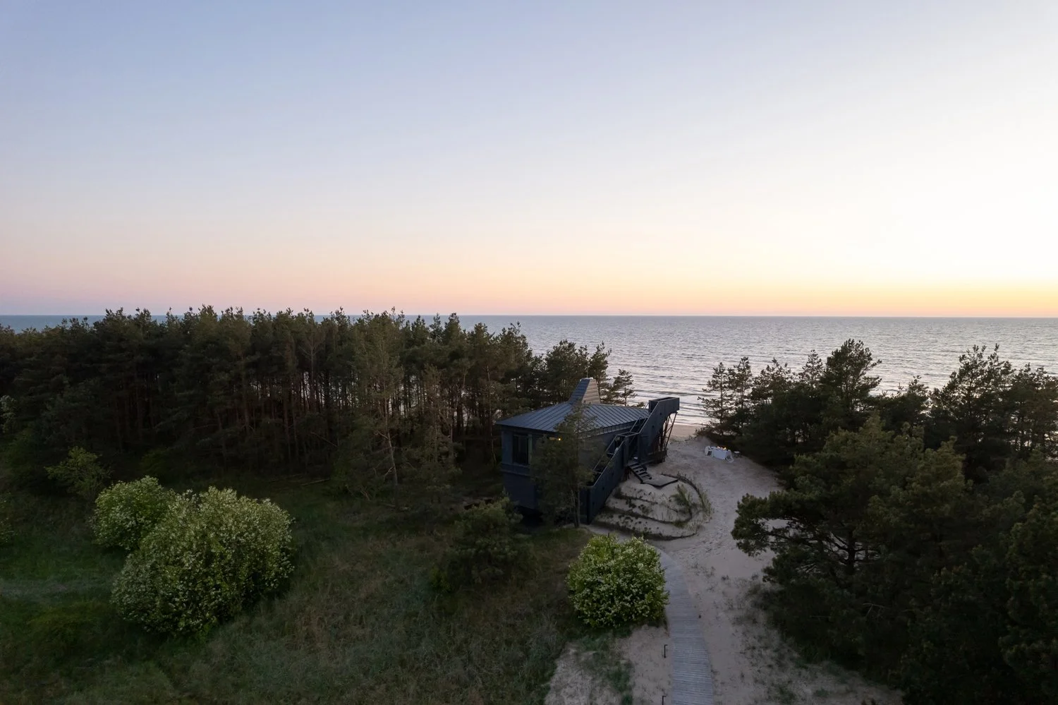 A coastal scene at sunset with a small beach house surrounded by trees and the ocean in the background.