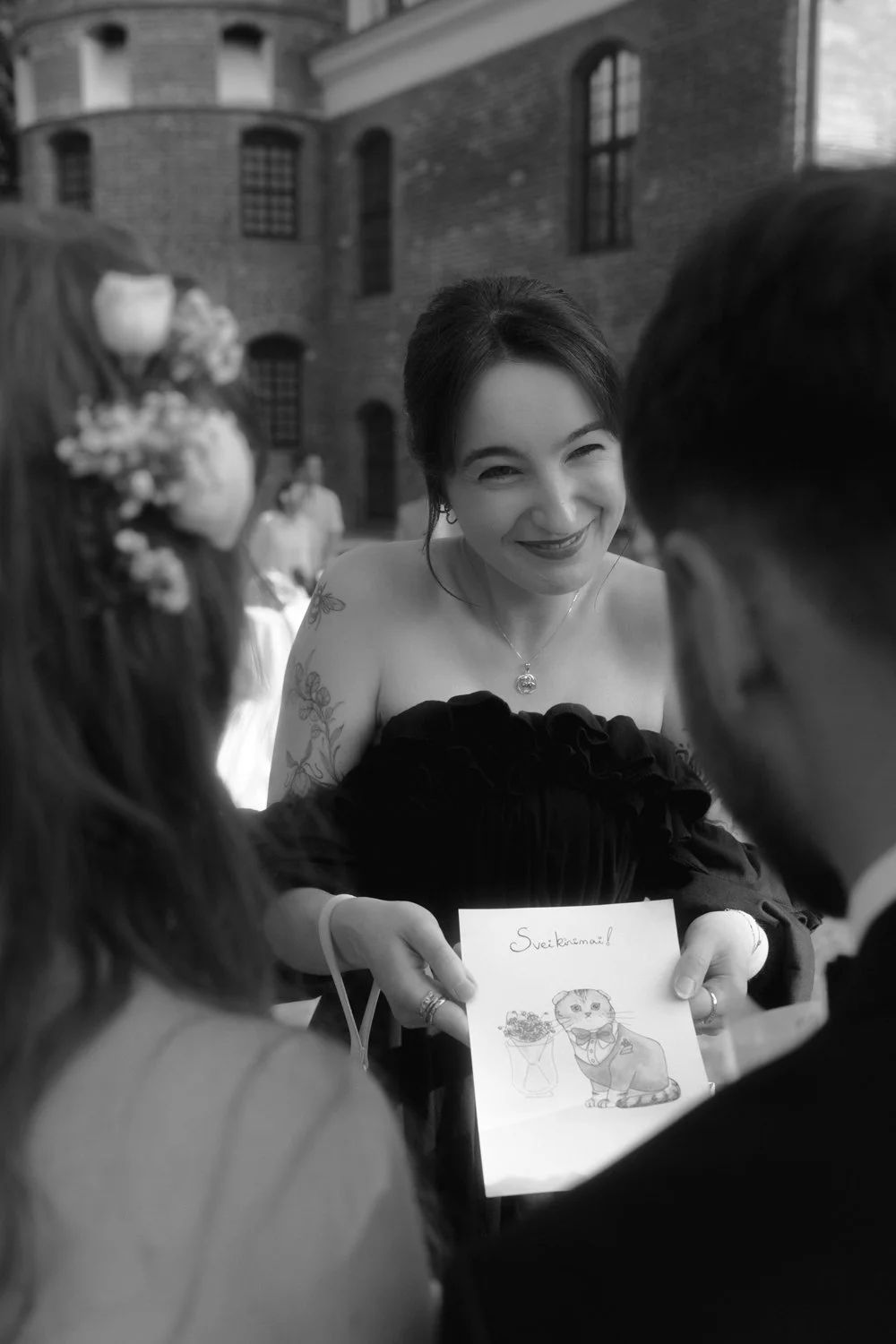 A woman smiling and showing a drawing of a cat wearing a bowtie, holding a cup, with the word 'Sviėkinamai!' written above, during a social event outside, with a brick building in the background.