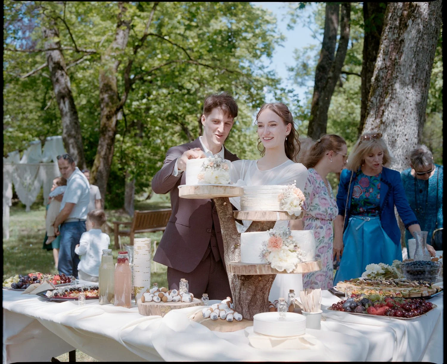 People celebrating a wedding outdoors, with a table of desserts and a couple cutting a wedding cake.