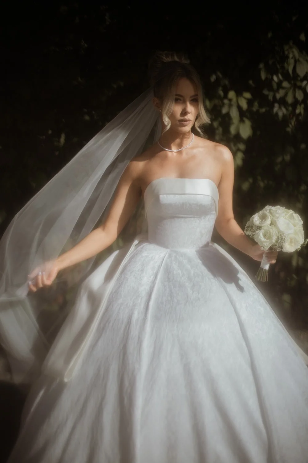 A woman in a strapless white wedding gown holding a bouquet of white roses, standing outdoors with dark foliage in the background.