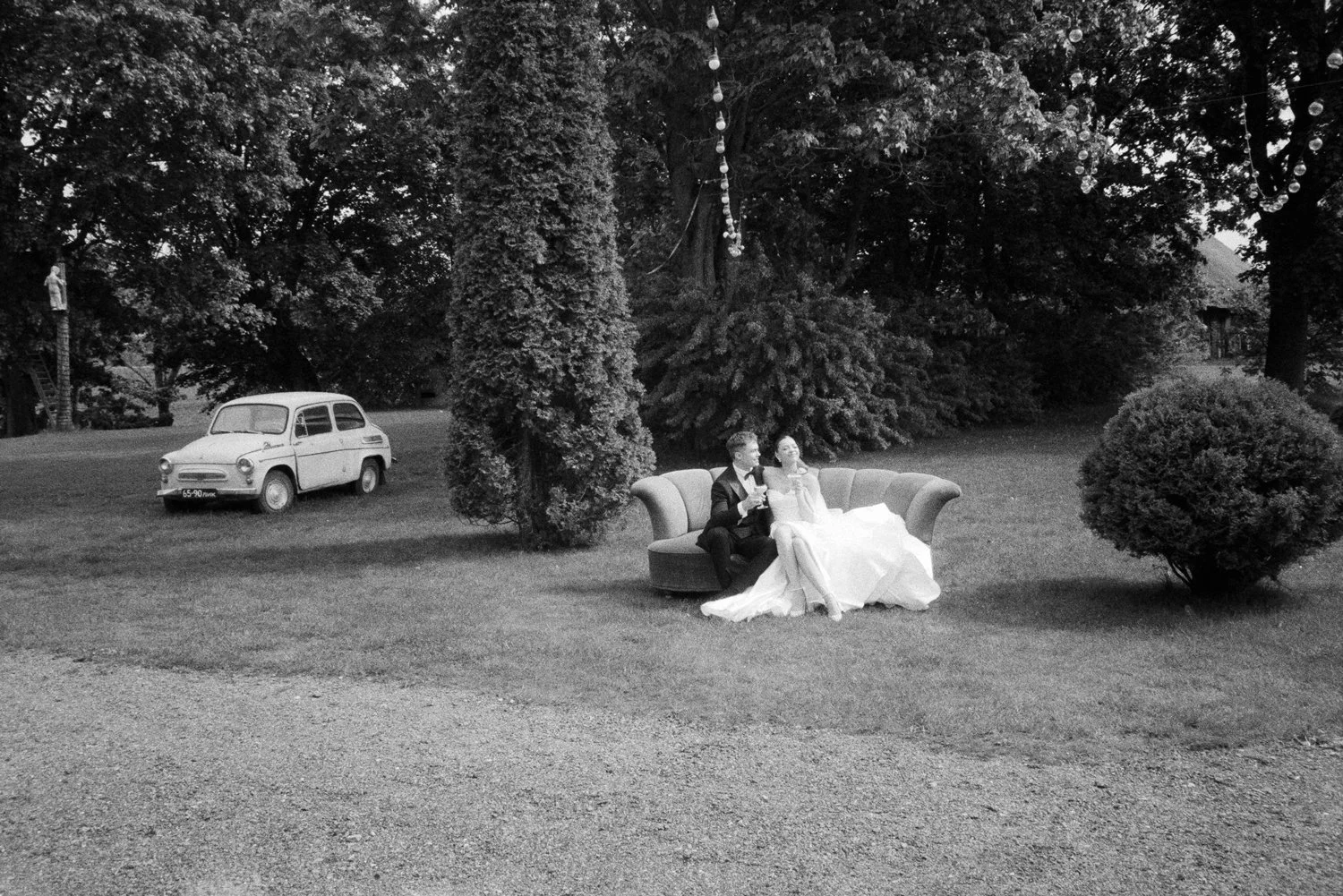Black and white outdoor scene with a vintage car on the left and a couple dressed in wedding attire sitting on a sofa in the middle, surrounded by trees and decorative lights hanging above.